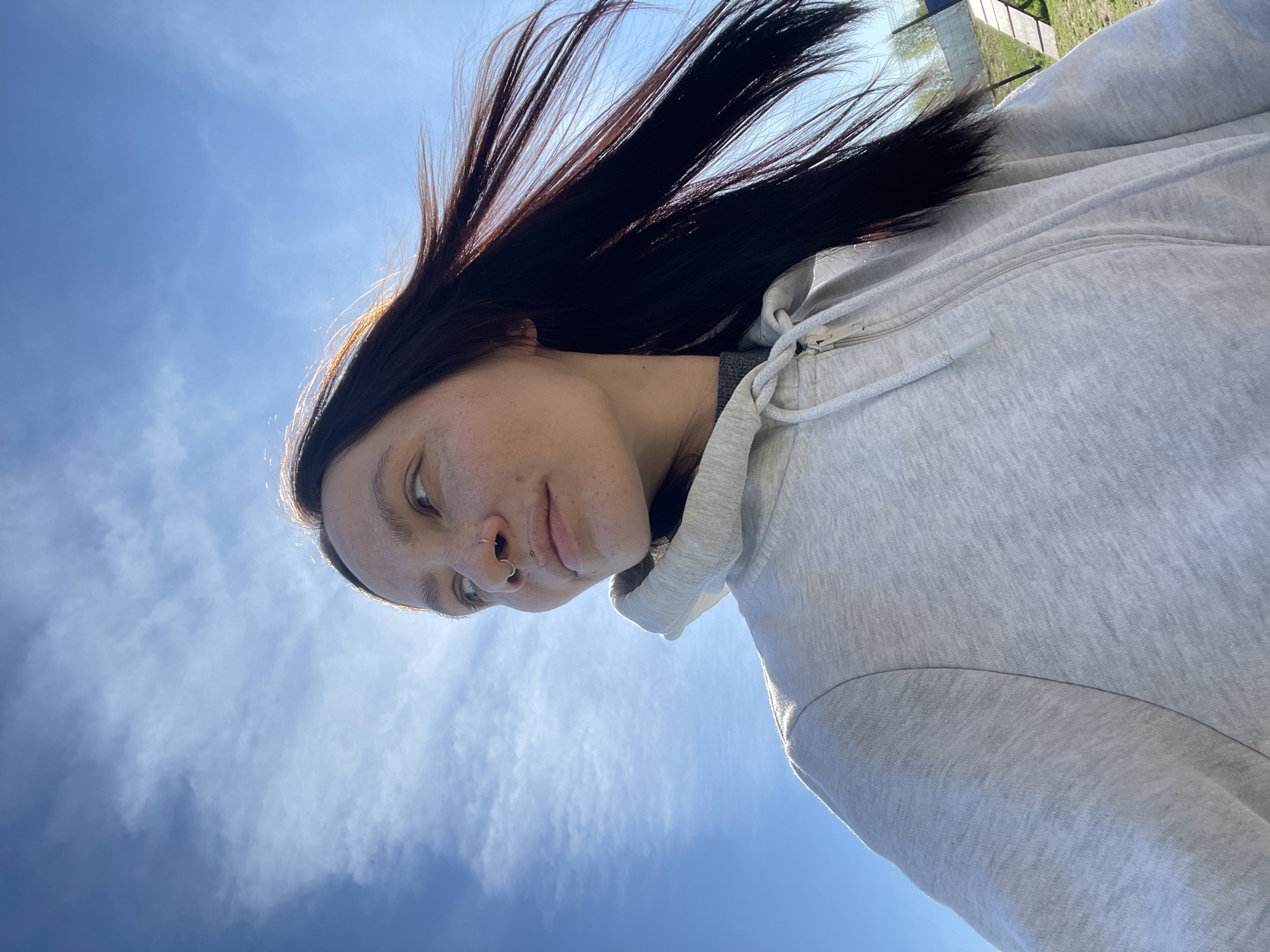 A low-angle, upward-facing photograph shows a person with dark hair wearing a light gray hooded sweatshirt. The person has a nose ring and is looking down and to the left. The background is a blue sky with wispy clouds. Faintly visible in the upper right corner is a green area, possibly grass, and a blurred structure that might be a fence or building. There's no information available to determine if the image was taken in Pervomaysky, Russia.