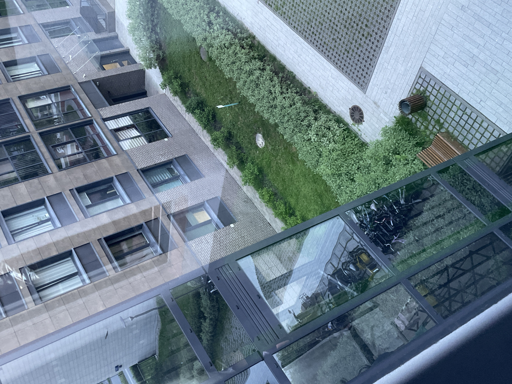 This image captures a view of a modern apartment building in Sinyavino, Russia, likely from an elevated vantage point. The building features a facade of light brown and dark grey materials, with numerous windows. The windows reflect the surrounding environment, including green foliage and portions of other buildings.
In the foreground, a reflection in a glass-covered area shows several bicycles and what appears to be storage or equipment. Below this, a wooden bench sits near a trash receptacle. The ground is paved with light-colored rectangular tiles and in some areas, a grid pattern of permeable pavers with grass growing through them. A section of meticulously maintained green bushes and grass is visible adjacent to the paved areas, suggesting a well-kept courtyard or common space. A manhole cover is also present in the paved section. The overall impression is of a contemporary residential area with designed outdoor spaces.