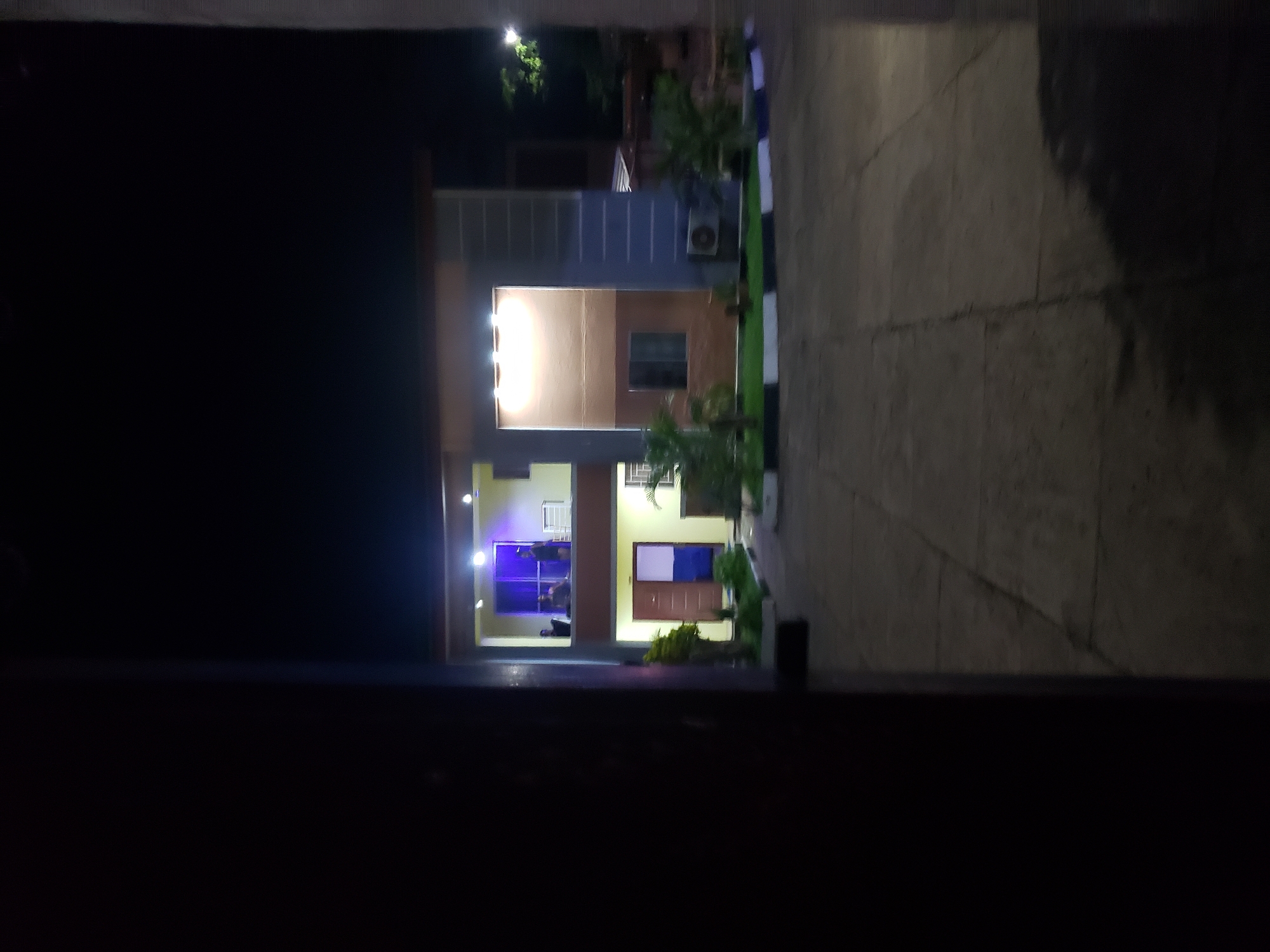 A nighttime exterior shot captures a modern two-story building in Narayi, Nigeria. The building features a flat roof, concrete facade, and balconies. The ground floor has an open doorway with a blue and white section inside. The second floor has a balcony with several people visible, illuminated by purple lighting. Landscaping with small trees and decorative borders lines the paved driveway. An air conditioning unit is mounted on the side of the building. The scene is viewed from behind a dark, vertical foreground object, possibly a doorway or pillar, partially obscuring the left side of the image.