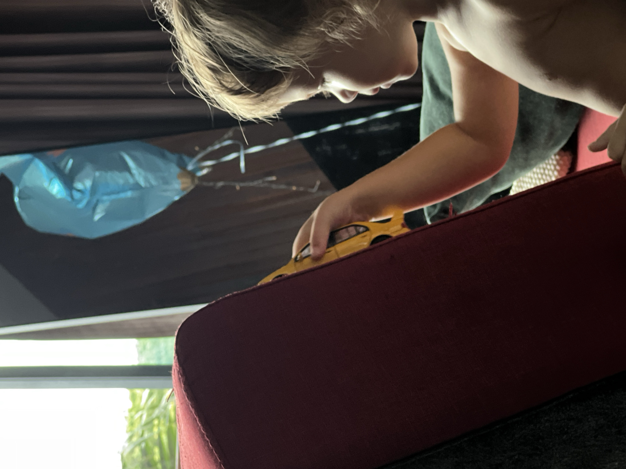 A young child, facing away from the camera, plays with a yellow toy car on a dark red sofa. The child has fair hair and is shirtless. To the left of the sofa is a window with a view of green foliage. In the background, dark curtains hang, and a blue balloon is visible. The scene takes place indoors, likely in a residential setting.