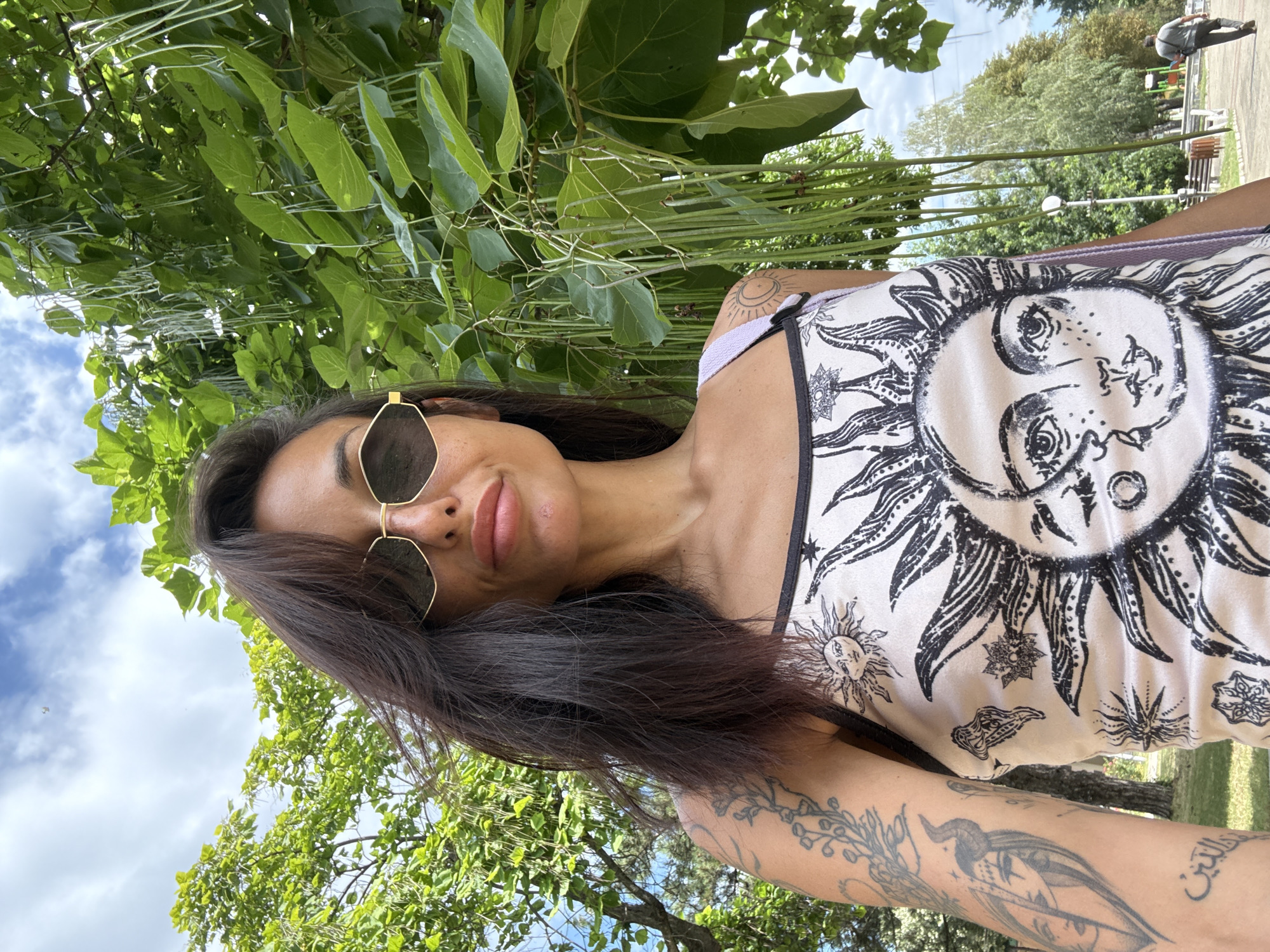 In the city of Belene, Bulgaria, a woman with dark hair and sunglasses is taking a selfie outdoors. She is wearing a tank top with a sun graphic. Her arm, visible on the left side of the frame, is tattooed. The background features lush green trees with long seed pods hanging from them, and a cloudy sky. In the distance, a street lamp, benches, and a person walking are visible.