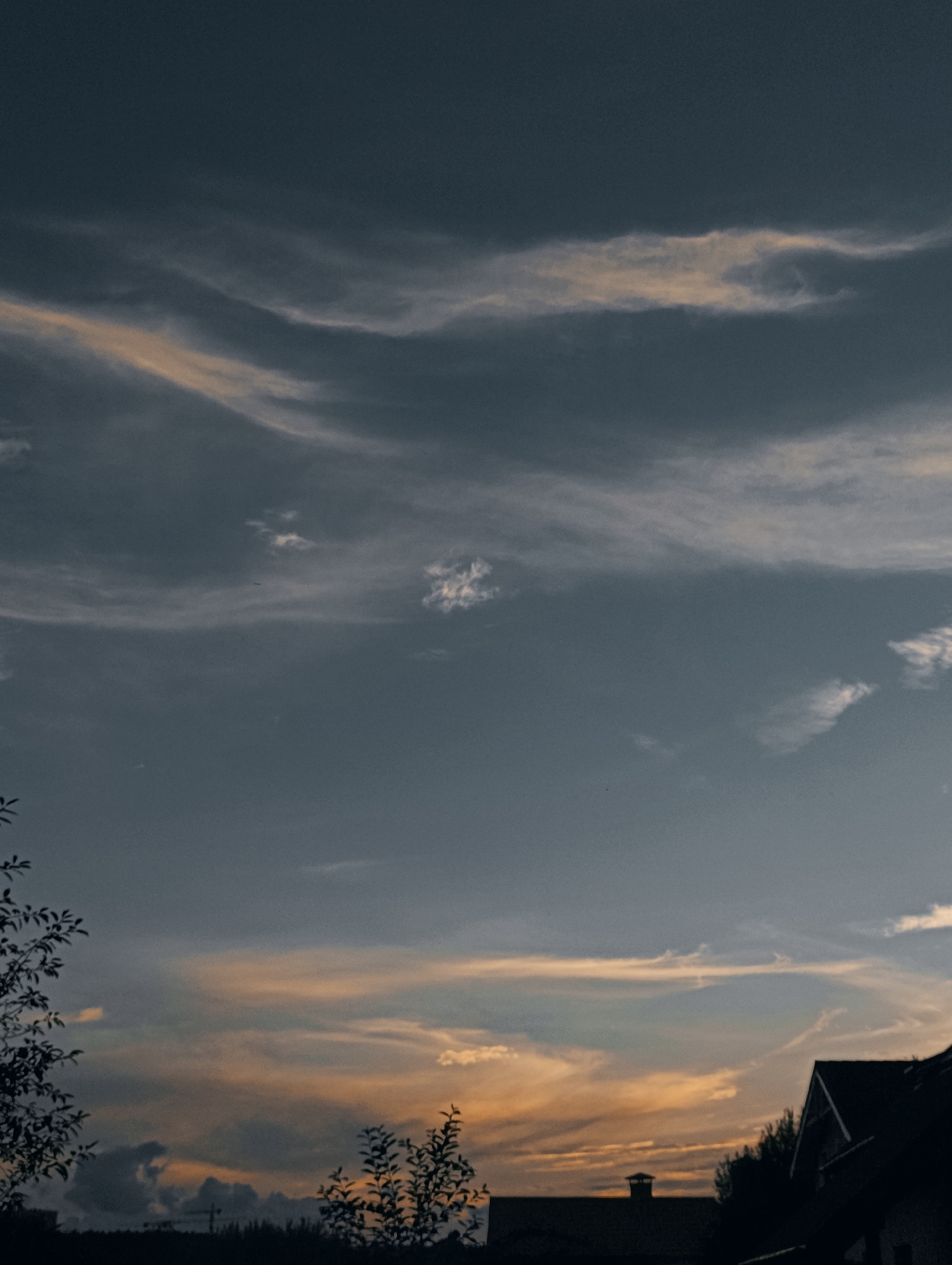 The image depicts a twilight sky with wispy, orange-tinted clouds interspersed with darker, bluish-gray clouds. The lower part of the frame shows the silhouettes of trees and rooftops, suggesting a residential area. A dark roofline with a gabled section and a chimney is visible on the right side of the image. The overall atmosphere is serene and slightly melancholic. The location is stated to be the city of Minsk, Belarus, and the scene likely captures a sunset or sunrise over a suburban or rural outskirts of the city. There are no people or animals visible in the frame.