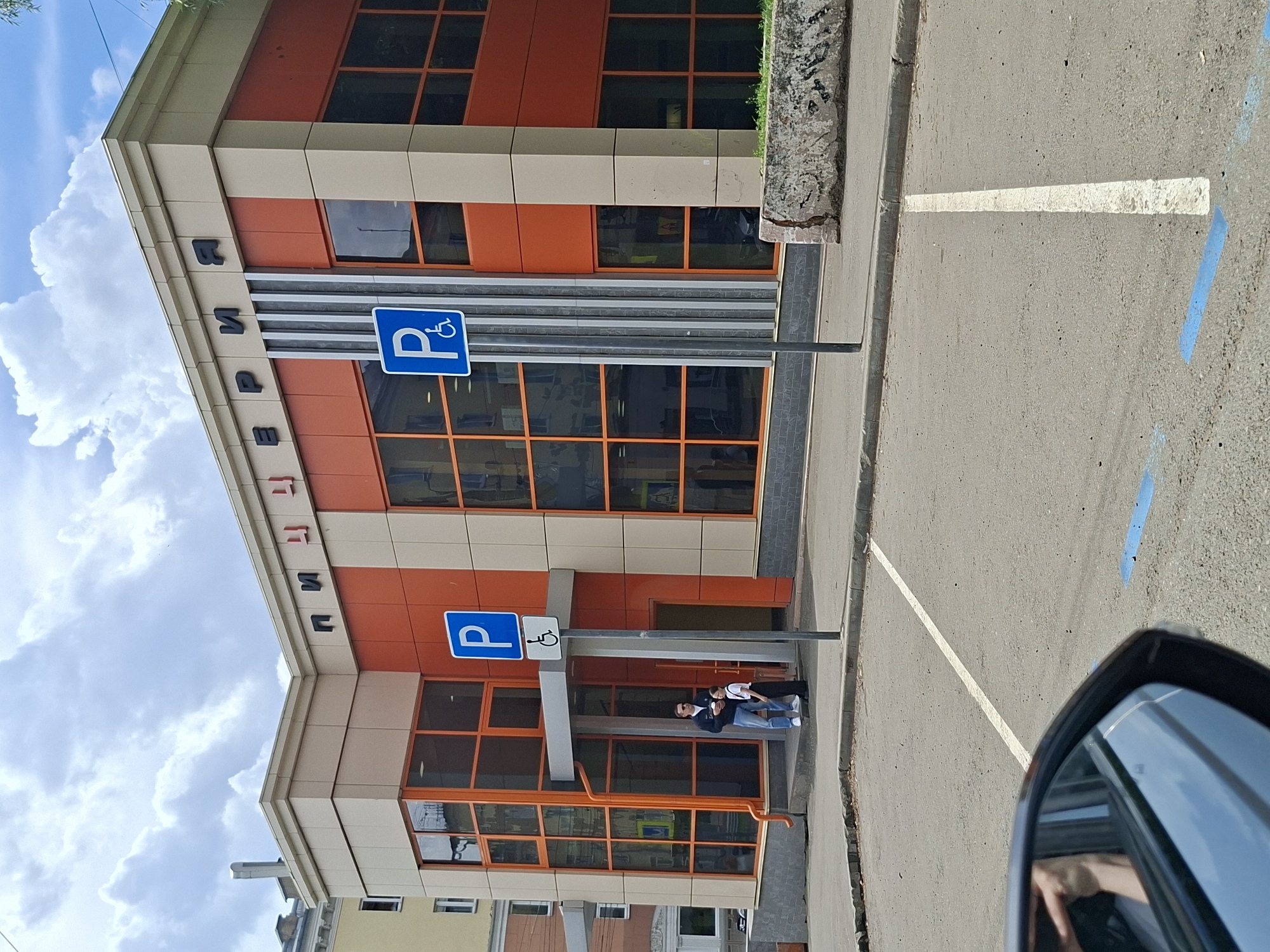 The image shows a street-level view of the exterior of a building with a sign reading "ПИЩЕРИЯ" (Pishcheriya) in black lettering. The building has a modern facade with large orange and beige panels, and large glass windows with orange frames. Two blue and white parking signs are visible, one with a wheelchair symbol indicating disabled parking. In front of the building entrance, a man holding a coffee cup stands with a young girl. A portion of a car's side mirror is visible in the foreground, reflecting a person's arm and hand. The scene is set on an asphalt surface with white parking lines and blue markings. The sky above is partly cloudy. This building is located in Izhevsk, Russia.