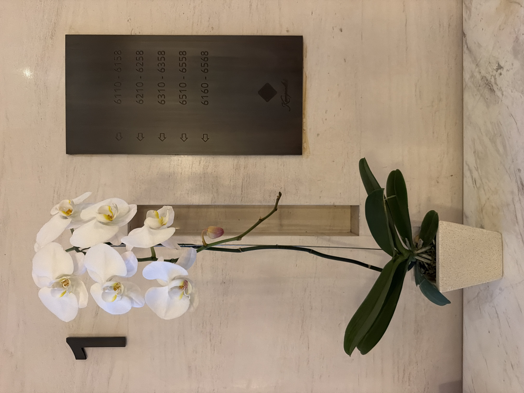 This image captures a detail of an indoor setting, likely a hotel hallway or lobby in Kuta Selatan, Indonesia. The main subjects are a potted white Phalaenopsis orchid and a dark rectangular sign mounted on a textured wall.
The orchid, situated on the left, features several pristine white blooms with yellow centers and a single pinkish bud, extending from a long green stem with broad, dark green leaves. It is planted in a light beige, textured square pot. A thin support wire or string is visible, seemingly holding the orchid stem upright and attached to a recessed wooden or light-colored panel behind it. Below the orchid blooms, a small, dark L-shaped decorative or functional object is attached to the wall.
Above the orchid, a prominent dark grey or black rectangular sign is mounted on the wall. This sign displays room number ranges vertically: "6110 - 6158", "6210 - 6258", "6310 - 6358", "6510 - 6558", and "6160 - 6568". Below each range, a small arrow points left. On the right side of the sign, there is a small diamond-shaped symbol and a stylized logo or signature that appears to read "Kempinski".
The walls consist of a light-colored, slightly textured material, possibly plaster or a fine stone finish, with a section of white marble with grey veining visible on the far right. The image conveys a sense of elegant and well-maintained interior space. There is no activity taking place, and no people are visible. The lighting is soft and even, typical of an indoor environment, making it impossible to determine the time of day.