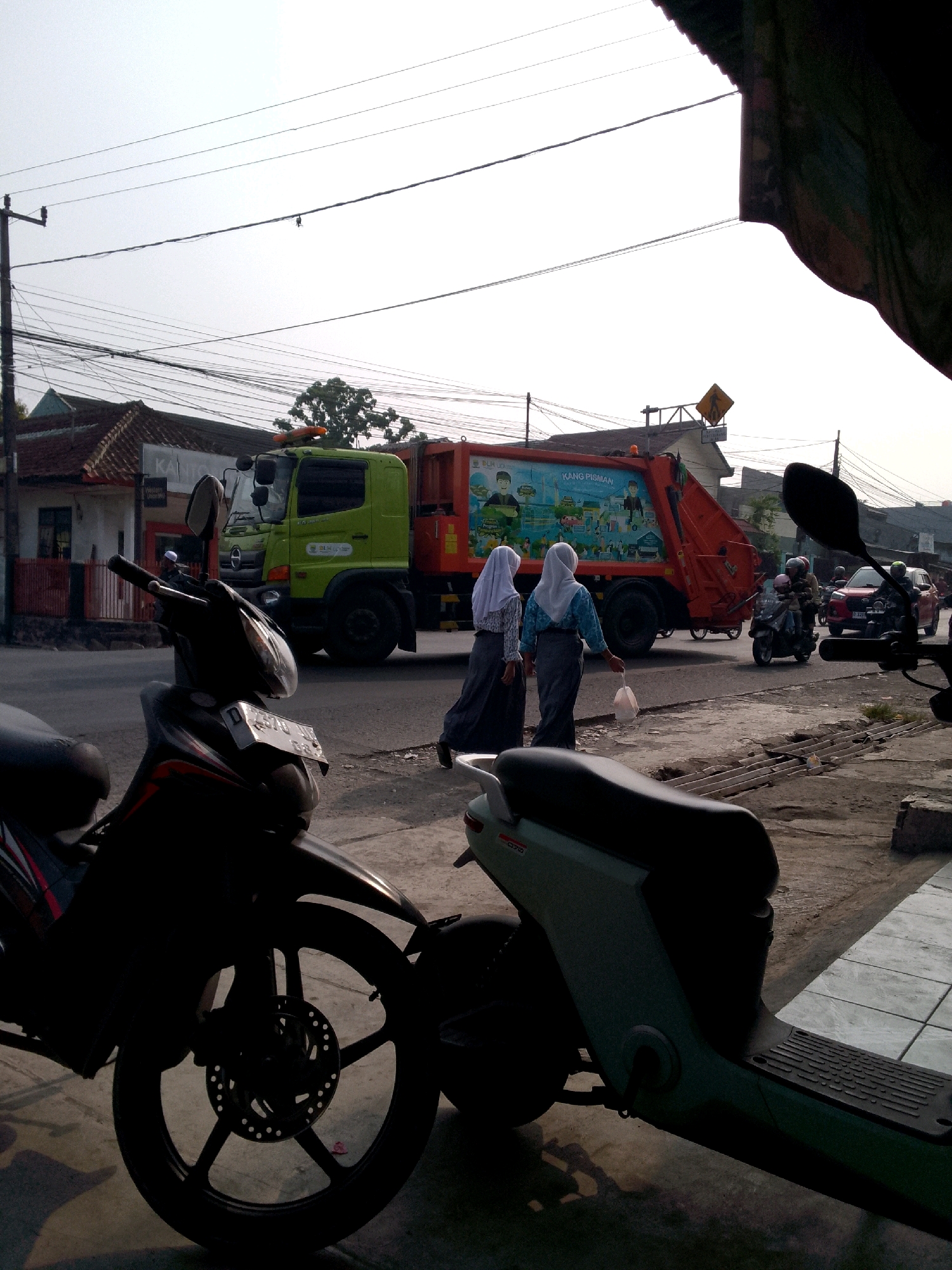 In Rajamandala Kulon, Indonesia, a garbage truck with an orange rear loading mechanism and a green cab is stopped on a street, adjacent to a sidewalk. Two young women in hijabs, one in blue and white, the other in purple and white, are walking near the truck. The woman on the right is carrying a white plastic bag. A dark-colored motorcycle is parked in the foreground, partially obscuring another light green electric scooter. Behind the garbage truck, traffic can be seen including a motorcycle with two riders and a red car. The scene is set against a backdrop of buildings, overhead power lines, and trees. The sky is overcast, suggesting it is daytime.