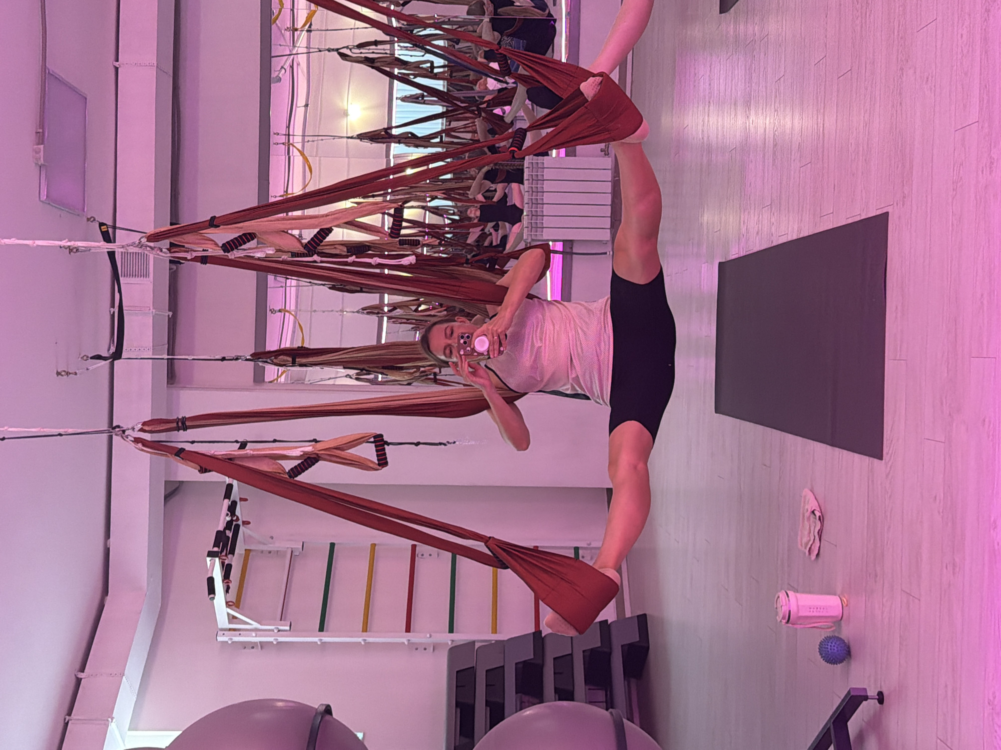 A woman is practicing aerial yoga in a studio, suspended in a split position by red yoga silks. She is holding a smartphone, taking a photo of herself. The studio has a mirrored wall, a wall-mounted pull-up bar, step platforms, and other yoga equipment. The lighting is predominantly pink.