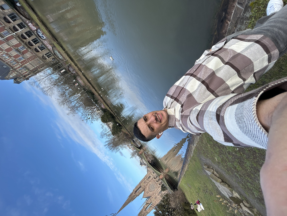 A male individual is depicted in a selfie-style photograph, standing on a grassy bank adjacent to a river in Strasbourg, France. The subject, who appears to be in his 20s or 30s, has short dark hair and light facial hair, and is wearing a brown, beige, and white plaid long-sleeved shirt, grey trousers, and white sneakers. White wireless earbuds are present in both ears as he looks towards the camera with a slight smile.
The location is identifiable by the prominent Gothic Revival Église Saint-Paul (Saint Paul's Church) with its twin spires, situated on an island in the background to the left. A green metal arch bridge (Pont Saint-Paul) is visible behind the church. The body of water is a river, likely the Ill River, which exhibits a calm, reflective surface with several birds floating upon it.
The immediate foreground features green grass and some bare earth, with scattered stones along the water's edge. In the far left background, two indistinct figures are seated on the grassy bank. The opposite riverbank displays bare deciduous trees and a multi-story building with reddish-brown brickwork and a dark roof, notable for graffiti marks along its base. The sky above is clear blue with sparse white clouds, indicating a bright, sunny daytime environment.