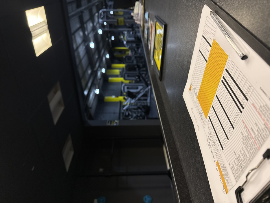 An interior image depicts a gym or fitness facility in Tunbridge Wells, United Kingdom, viewed from behind a dark, textured counter. In the foreground, a white paper schedule, dated "Thursday 19th March 202X," rests on a black clipboard with a silver pen. The schedule displays a grid format with a large yellow block and rows containing black horizontal lines, along with handwritten notes including "Have a great day!" and names like "Liv" and "Kim." Beyond the counter, the gym area is visible, featuring dark walls and ceiling, contrasting with bright yellow vertical support pillars. Numerous black and metallic exercise machines are arranged on the floor. Overhead lighting consists of multiple circular, recessed white lights and square ceiling lights. A dark wall to the left features several blue, circular objects resembling climbing holds. No individuals are clearly depicted.