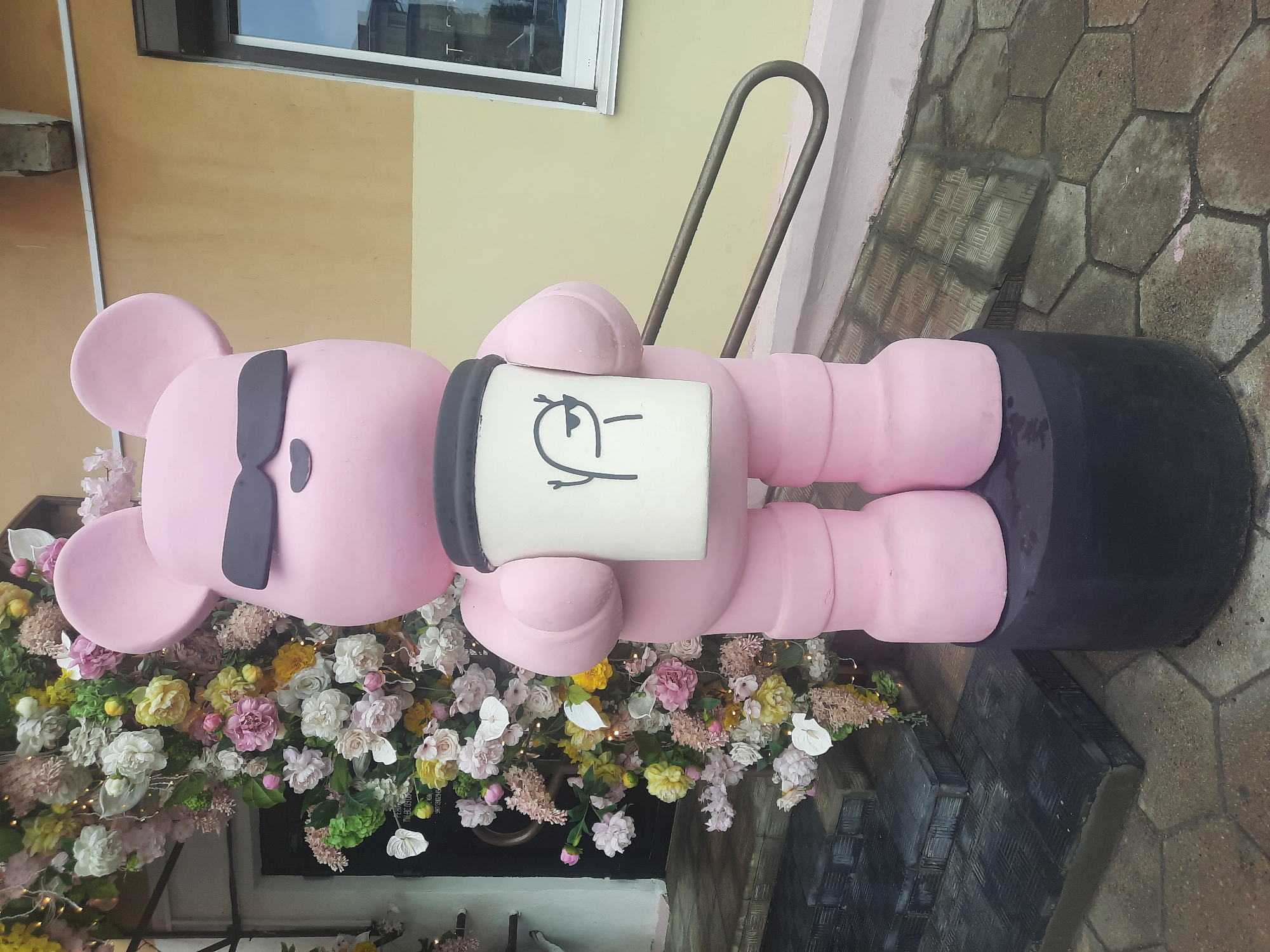 A large, pink, cartoon bear statue stands on a black pedestal outside a building with a yellow facade. The bear is wearing black sunglasses and holds a white coffee cup with a drawing of a person on it. To the left of the bear, there is a doorway framed by an archway of artificial flowers and string lights. In front of the doorway are dark, textured stairs. A metal handrail is visible to the right of the bear. The ground in front of the statue is paved with hexagonal stones. The image appears to be taken in the city of Hrodna, Belarus.
