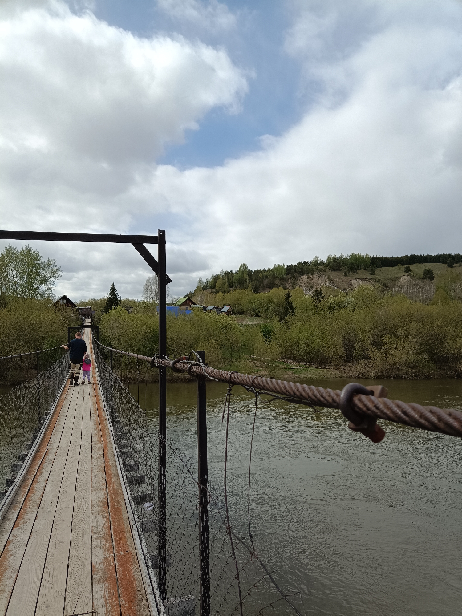 The image shows a suspension bridge spanning a river, likely located in or near Посад, Russia, given the landscape and architectural details. The bridge is constructed of wooden planks and metal cable railings with chain-link fencing on the sides.
A man and a young child are walking across the bridge, away from the camera. The man is wearing a dark shirt and shorts. The child, closer to the camera, is wearing a pink top and dark pants.
The river below is murky green in color. On the opposite side of the riverbank, there is dense foliage, including trees and bushes. In the background, there are structures that appear to be houses or cabins.
The sky is overcast with grey and white clouds, some with patches of blue sky. The bridge structure is visible in the foreground, with metal cables and supports.