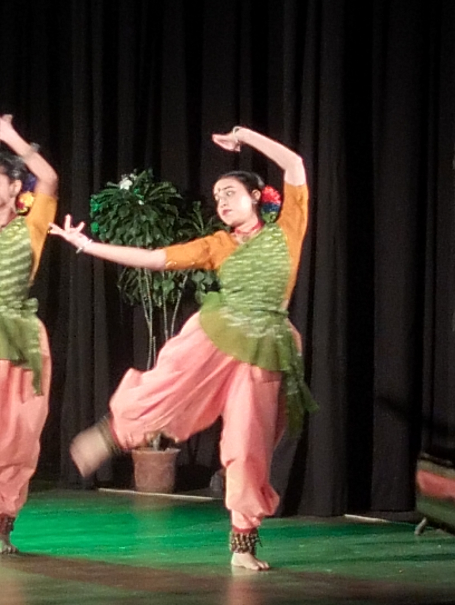 Two female dancers perform on a stage. The dancer in the foreground is wearing a mustard-yellow long-sleeved shirt, a green and white patterned vest, and loose-fitting pink pants. She has her left arm raised and bent at the elbow, with her hand open, and her right arm extended outwards. Her left leg is bent and her foot is raised, with ankle bells visible. The dancer on the left is partially visible and wears similar attire. They are positioned in front of a black curtain. In the background, a potted plant with green leaves is situated on a shelf or platform. The stage floor is illuminated with green lighting. The scene appears to be a theatrical performance.