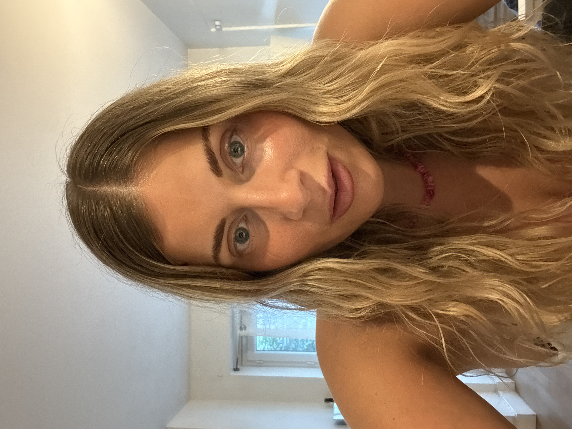 A light-skinned woman with wavy blonde hair is looking directly at the camera. She is wearing a pink beaded necklace. The background is a white wall with a window visible. The location cannot be determined to be Paris, France from the visual information provided.