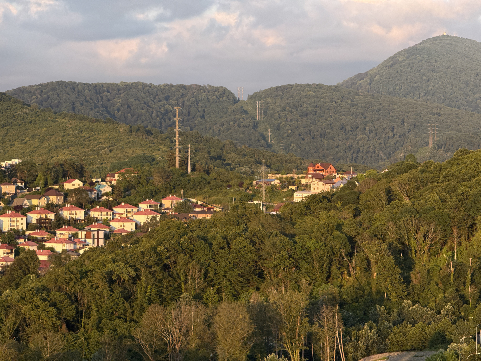 This is a high-angle, wide shot photograph of a densely forested hillside in Sochi, Russia, dotted with residential buildings. In the foreground, a thick canopy of green trees dominates the lower portion of the frame, with some bare trees visible in the undergrowth. A dirt path curves up from the bottom right corner.
The middle ground reveals a sprawling residential area composed of numerous houses with red-tiled roofs and white or light-colored walls. The buildings are clustered together on the undulating terrain, suggesting a suburban or semi-rural setting. Power lines and transmission towers are visible throughout the landscape, indicating the presence of infrastructure.
In the background, lush green mountains rise, their slopes covered in dense forest. Several transmission towers are strategically placed on the mountainsides and in the valleys between them, with a prominent tower on the left and a series of towers cutting through the gap between two peaks. The sky above is a pale blue with scattered clouds, tinged with pink and orange from what appears to be a sunset or sunrise. The overall impression is one of a developed landscape integrated with a natural, mountainous environment, likely in a coastal city like Sochi known for its subtropical climate and mountainous terrain.