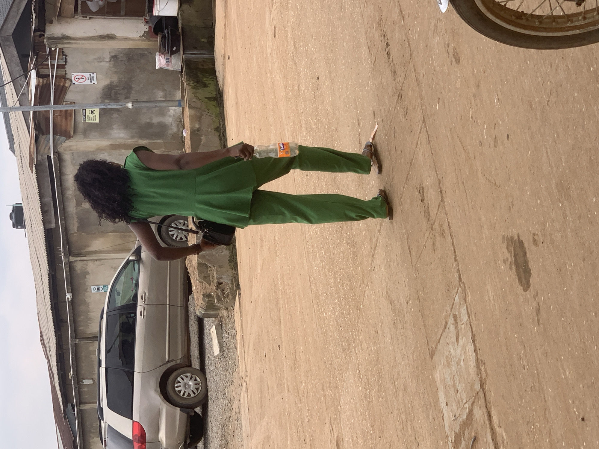 A woman with dark, curly hair, seen from behind, walks on a dirt road in Shagamu, Nigeria. She is wearing a green, sleeveless peplum top and matching green pants, along with brown sandals. In her right hand, she carries an open bottle of Fanta. She is also carrying a black purse on her left shoulder. To the left of the woman, a beige minivan is parked. The background features a concrete wall with various signs and a corrugated metal roof. The ground is covered in reddish-brown dirt with scattered debris and some paved sections. The sky is overcast.