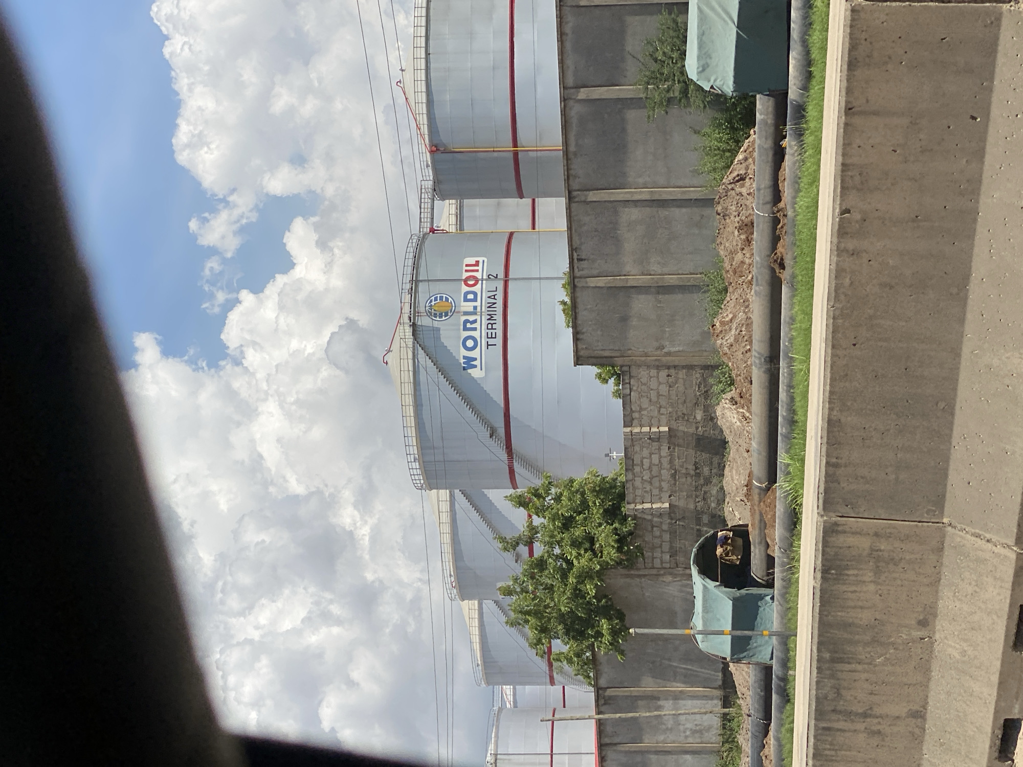 The image shows several large, cylindrical storage tanks with a white finish, typical of those used for petroleum or similar products. The tanks are labeled "WORLDOIL TERMINAL 2".
The tanks are situated behind a tall concrete barrier or wall. Adjacent to the wall, there's a small area of unkempt vegetation and exposed soil, with some pipes running along the barrier.
The sky above the tanks is partly cloudy with a mix of blue sky and puffy white clouds. The angle of the shot suggests it was taken from a vehicle, with a portion of the vehicle’s interior visible in the corner of the frame.
Based on the label "WORLDOIL TERMINAL 2," the location is likely within the city of Dar es-Salaam, Tanzania, where such industrial facilities are often found.