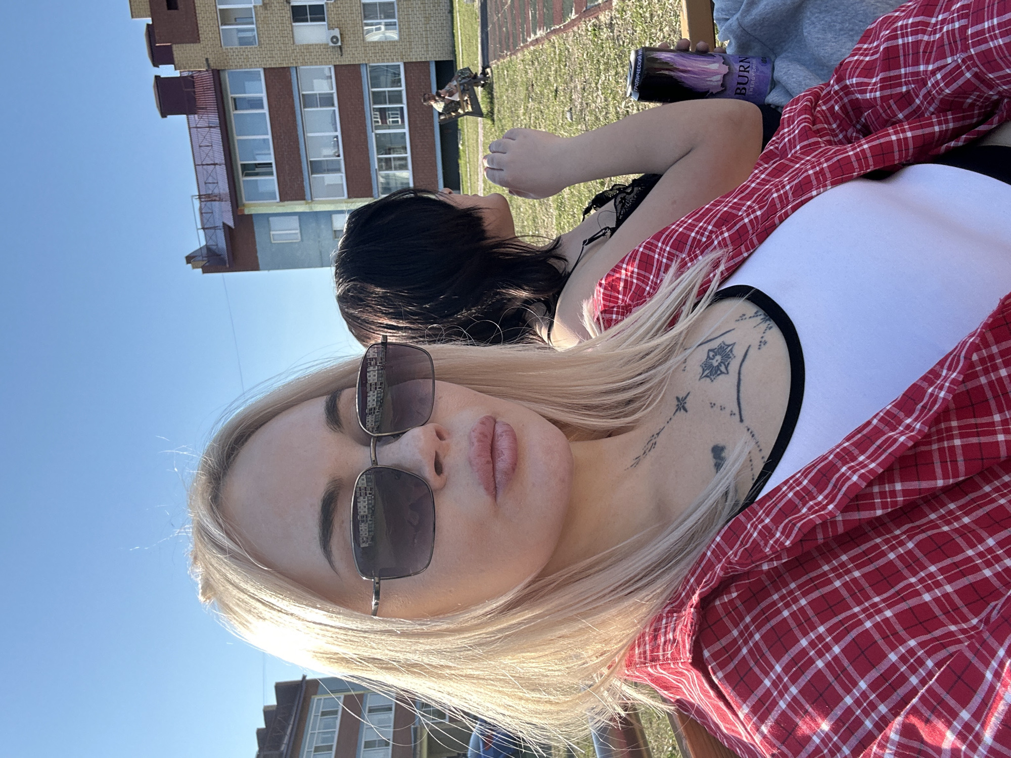 A daytime outdoor shot taken in Высокий, Russia, features a blonde woman wearing sunglasses and a red plaid shirt over a white tank top. Tattoos are visible on her chest and neck. Behind her, a woman with dark hair is partially visible, holding a can of "BURN" energy drink. In the background, there is a building and figures of two people sitting on a bench in a park area.