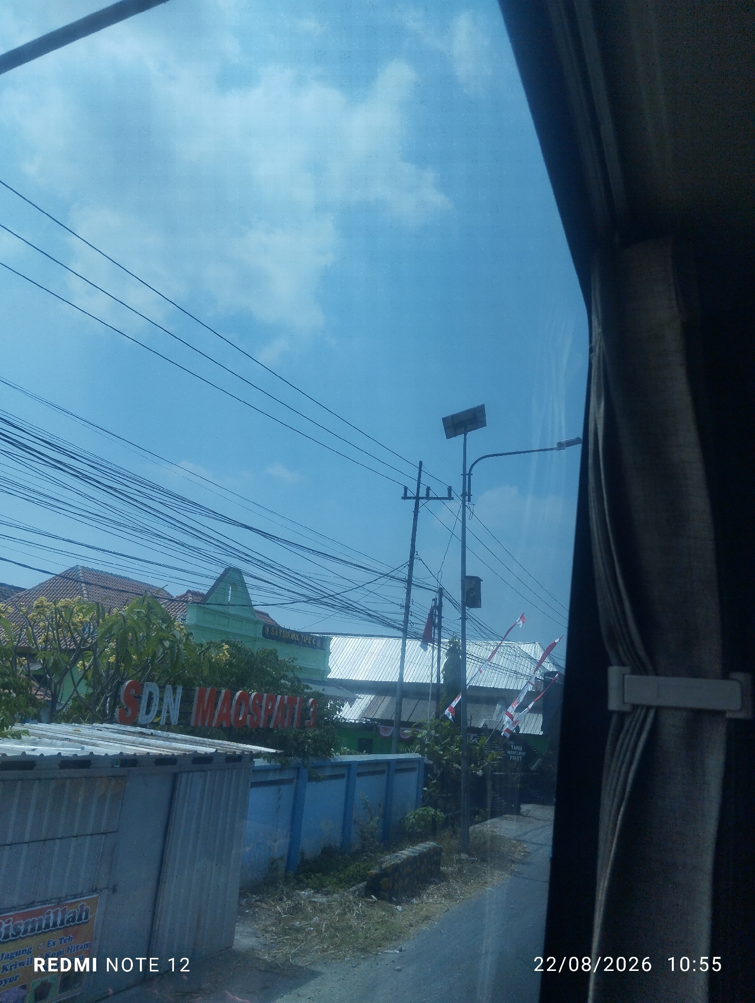 The image depicts a street scene in Kraton, Indonesia, with a clear blue sky and scattered clouds. A large number of electrical wires crisscross the upper portion of the frame, suggesting an urban or semi-urban environment. In the mid-ground, a green building with a red-tiled roof is visible, displaying signage that reads "SDN MAOSPATEL." This indicates the presence of an elementary school. To the left of the school is a grey metal shed or structure with some writing on it, partially obscured. A blue wall runs along the property line. A street lamp with a solar panel is also visible, along with some Indonesian flags. The right side of the image is dominated by a dark curtain or drapery, likely from a vehicle's window. The timestamp "22/08/2026 10:55" and the text "REDMI NOTE 12" are overlaid on the lower portion of the image, indicating the device and date/time the photo was taken. The ground appears to be a paved road and unkempt grassy areas.
