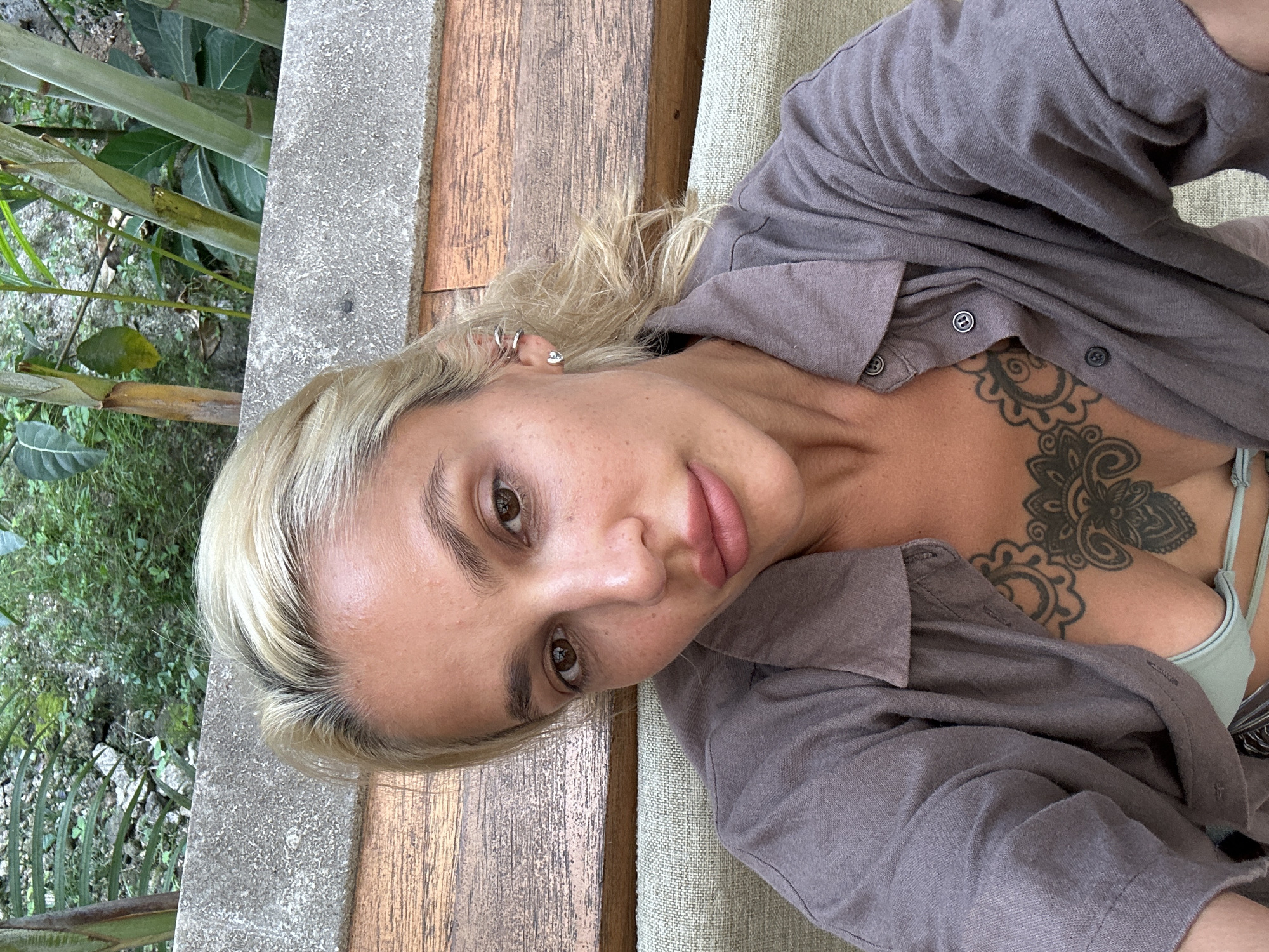 A medium-shot selfie shows a woman with blonde hair and light skin sitting outdoors. She is wearing a gray button-up shirt, open to reveal a light green bikini top and a floral tattoo on her chest. She has a silver heart earring in her left ear and three silver ear cuffs on her right ear. Behind her, a wooden bench and a concrete barrier are visible, with lush green foliage in the background, suggesting a tropical or garden setting.