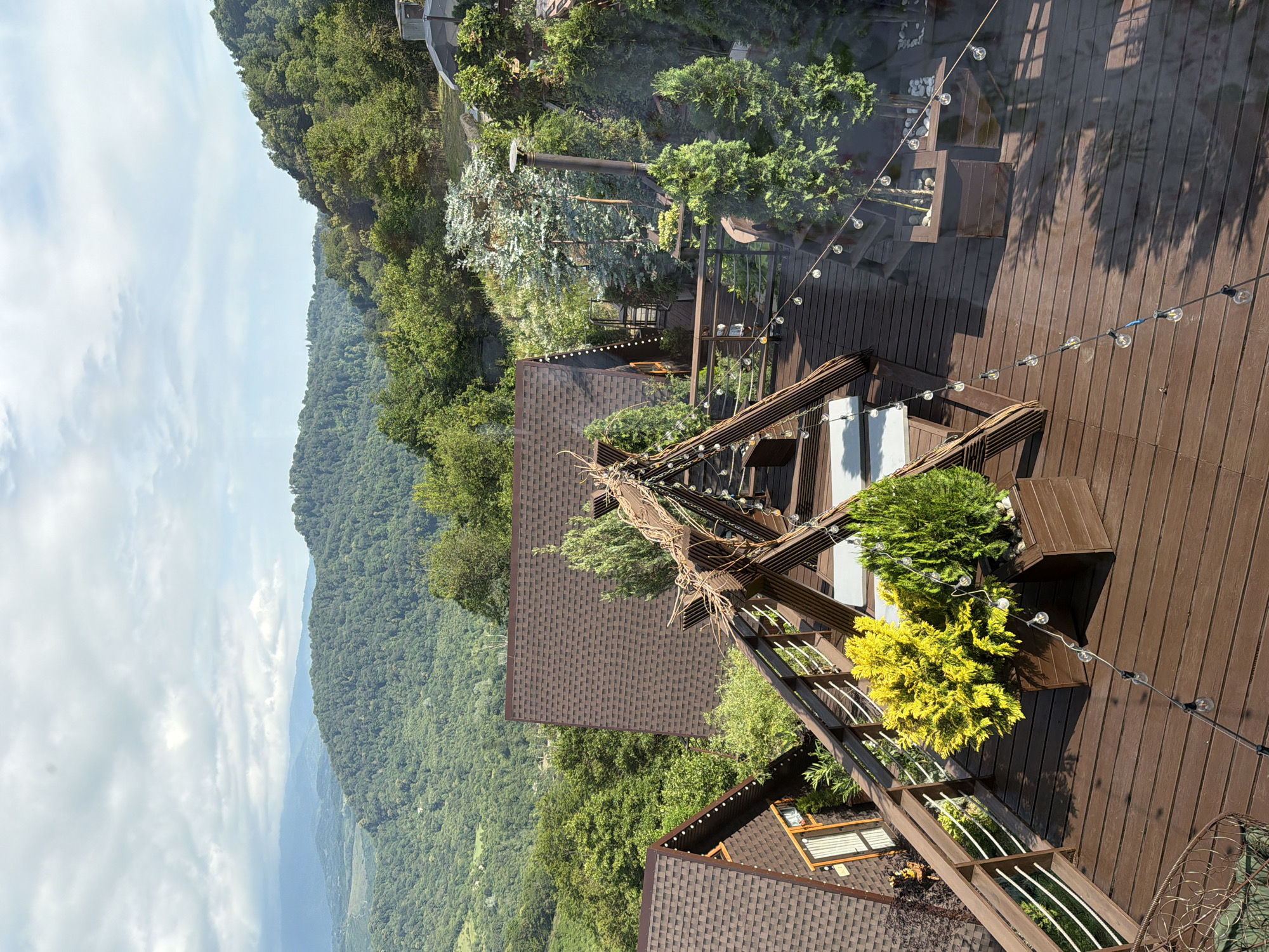 This is an eye-level, slightly tilted, exterior shot taken on a sunny day. The scene features a wooden deck with various decorative elements, including a string of lights and planters containing green and yellow foliage. A portion of a dark-tiled roof is visible in the foreground. The deck appears to be part of a building or establishment situated on a mountainside. Beyond the deck, a dense, verdant forest covers the undulating terrain, extending into the distance where it meets a hazy, pale blue sky with scattered clouds. The overall impression is one of a serene, natural landscape with man-made additions designed for outdoor enjoyment. There is no visible text or human activity. The location is identified as Nizhnyaya Shilovka, Russia.