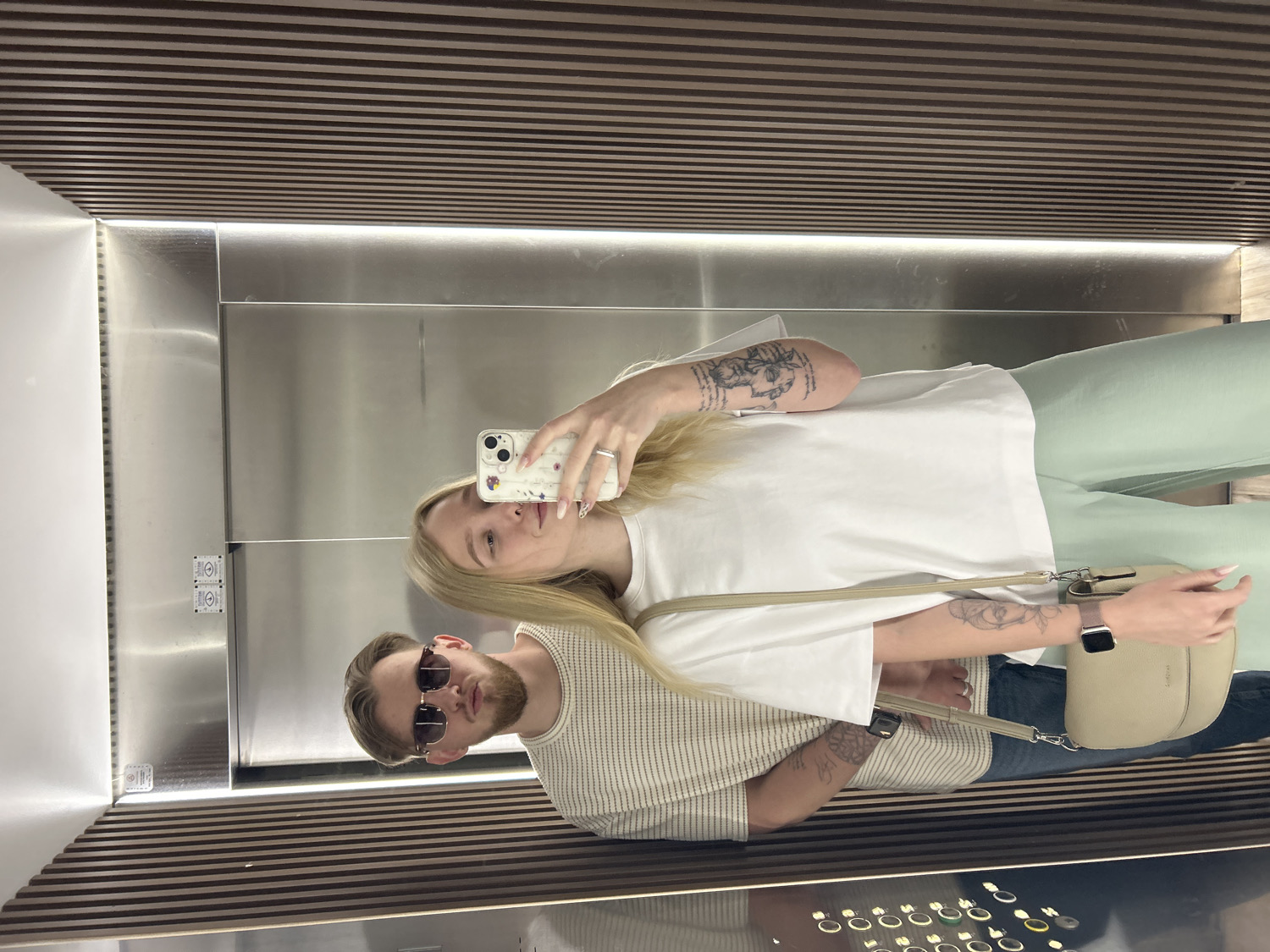 A mirror selfie captures two individuals, a woman and a man, inside what appears to be a modern elevator, likely in Budva, Montenegro. The image is oriented vertically, showing the ceiling and floor with horizontal wooden slat paneling, while the walls are a mix of reflective mirror panels and brushed metallic surfaces. Linear LED lighting illuminates the space from above the metallic wall panels.
The woman, with long blonde hair, is positioned on the left side of the frame. She wears a white oversized t-shirt and light green wide-leg pants. Her arms are adorned with tattoos, including what appears to be a classical bust on her upper left arm and a floral design on her lower right arm. She holds a white smartphone with a decorative case in her right hand, actively taking the selfie, and wears a ring on her left ring finger. A light-colored crossbody bag hangs from her shoulder, and a smartwatch is visible on her left wrist.
Beside her, on the right, stands a man with short brown hair and a beard. He is wearing dark aviator-style sunglasses and a light-colored, possibly striped or ribbed, top. Both individuals have neutral to relaxed expressions, with the woman's gaze directed towards the phone.
An elevator control panel is visible on the wall to the left of the subjects, featuring small pictorial signs (such as up/down arrows and a bell or phone symbol) and several circular buttons. No clear, legible text is visible on the panel, clothing, or accessories, though an illegible brand name might be present on the woman's bag. The scene depicts a casual moment captured in a contemporary indoor setting.