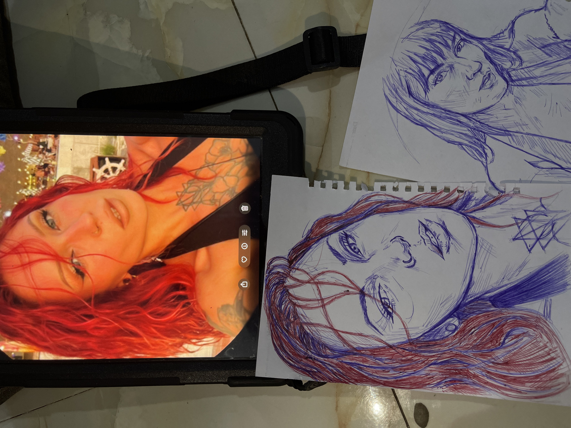 The image shows a tablet displaying a photograph of a woman with bright red hair and tattoos on her chest. Below the tablet are two sketches of women's faces, drawn on paper with blue and red ink. One sketch is larger and more detailed, showing the woman from the photograph's upper body. The other sketch is smaller and less detailed, depicting a different woman. The scene appears to be indoors, with a patterned floor visible. There is no indication of the city of Abuja, Nigeria, or any other specific location.