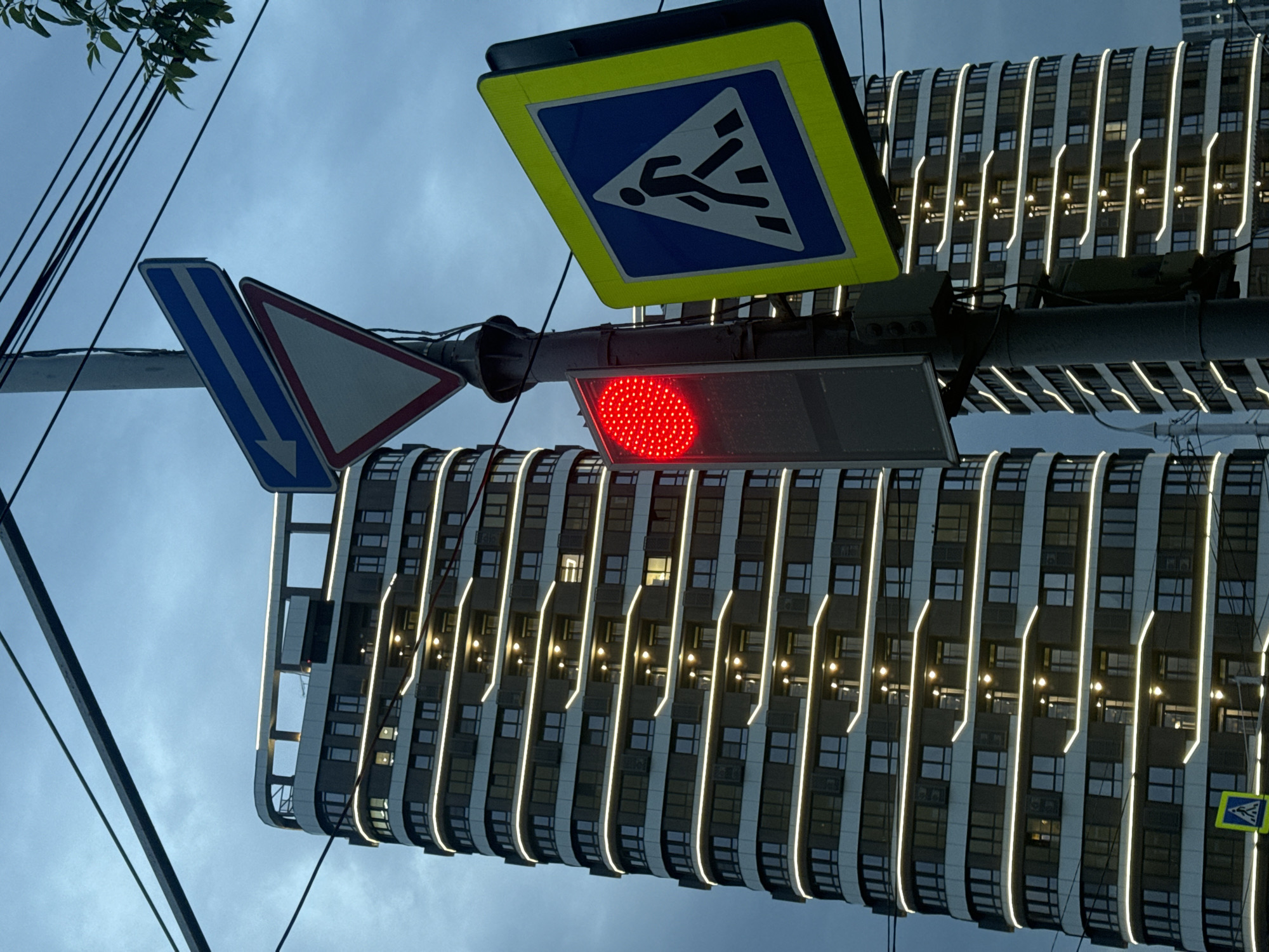 The image depicts a street scene in Krasnoyarsk, Russia, at dusk. A tall, modern, curved apartment building with illuminated horizontal bands of light is visible in the background. In the foreground, a traffic light pole displays a red traffic signal, a blue and white pedestrian crossing sign, and two other traffic signs: a triangular "yield" sign and a rectangular blue sign with white arrows indicating a left turn is mandatory. Overhead power lines cross the frame. The sky is overcast and a dark blue-gray, suggesting impending rain or evening. No people are visible in the image.
