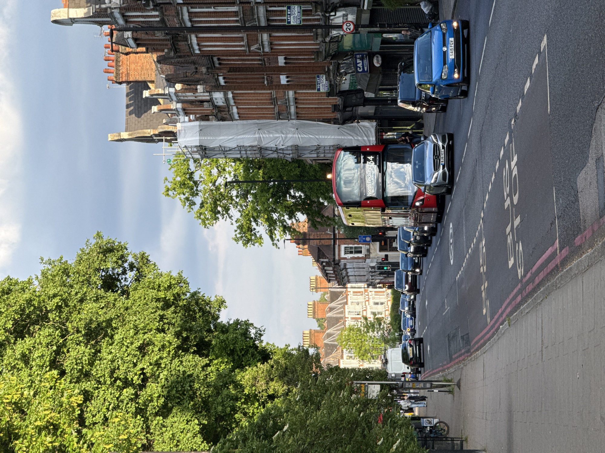 This is a street scene in London, United Kingdom, on a street with red brick buildings. Vehicles are lining the street, including a red double-decker bus and cars such as a blue Volkswagen. There are green trees on the left side of the image along the sidewalk. The image also includes details such as "North Finchley 134" on the front of the bus, a "Cafe Open" sign, "To Let" signs, and license plates, which provide contextual information. Road markings are visible on the asphalt. Scaffolding with white covering obscures part of one of the buildings.