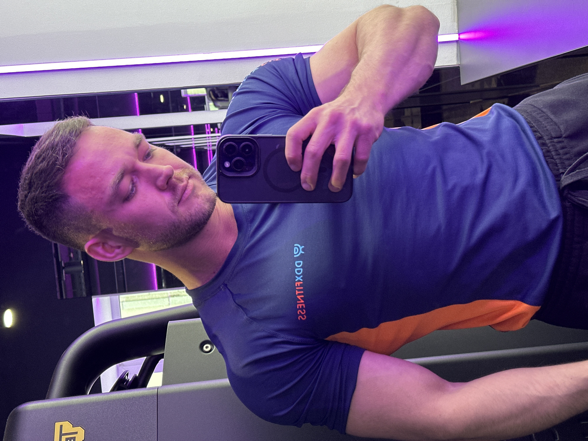 A man is taking a selfie in a gym. He is holding a black iPhone in his right hand, which is positioned in front of his face. The man is wearing a blue and orange athletic t-shirt. The gym has a purple-lit environment, with fitness equipment visible in the background. The text on the t-shirt reads "DDX FITNESS" in reverse. The city of Perm, Russia, is not visually verifiable from this image.
