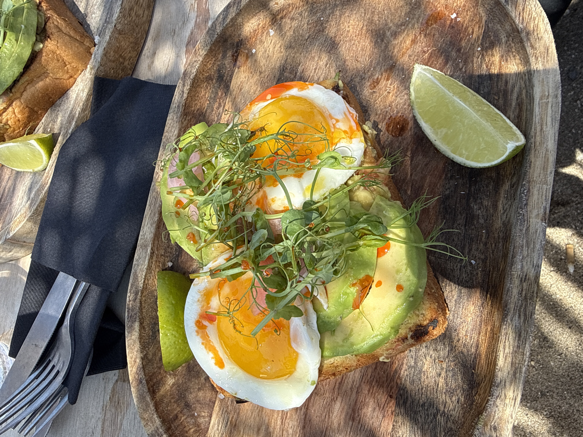 This is a close-up, overhead shot of an avocado toast with two fried eggs served on a rustic wooden board. The toast is topped with sliced avocado, two perfectly cooked fried eggs with runny yolks, and a garnish of fresh microgreens. A slice of lime is placed to the side of the toast, and another lime wedge is visible to the left of the frame. A dark napkin and a fork and knife are also present on the board, suggesting the meal is ready to be eaten. The background shows a sandy surface and the dappled sunlight indicates it is daytime, likely outdoors. The overall impression is a healthy and appealing breakfast or brunch. The setting appears to be a casual outdoor dining area, potentially a beach cafe, given the sand. The sunlight casting shadows on the wooden board suggests a sunny day. There is no visible text in the image.