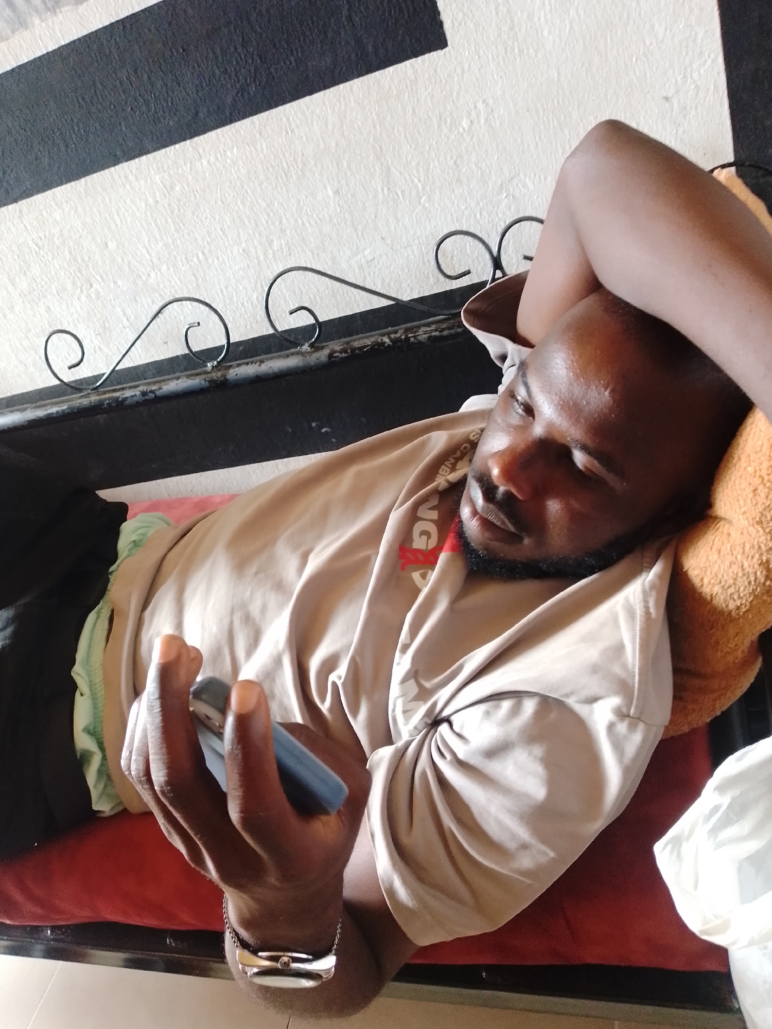 A man is depicted lying down, appearing relaxed and engrossed in a smartphone. He has dark skin, a short beard, and is resting his head on an orange-brown pillow. One arm is bent with his hand behind his head, while the other arm holds a dark-colored smartphone with both hands, suggesting he is actively using it. He is wearing a light brown/tan t-shirt that features a red graphic on the chest, where partial text like "G" and "AND" can be seen. A silver-colored bracelet or watch is visible on his left wrist.
The man is positioned on a red mattress or cushion. Behind him is a black metal frame, possibly part of a bed, adorned with decorative scroll patterns. The wall in the background displays a distinctive pattern of broad diagonal black and white stripes. Various fabrics, including green on the left and white on the right, are visible around him. The lighting indicates it is daytime, with soft highlights on his skin. The scene suggests an indoor setting, likely a bedroom or living area.