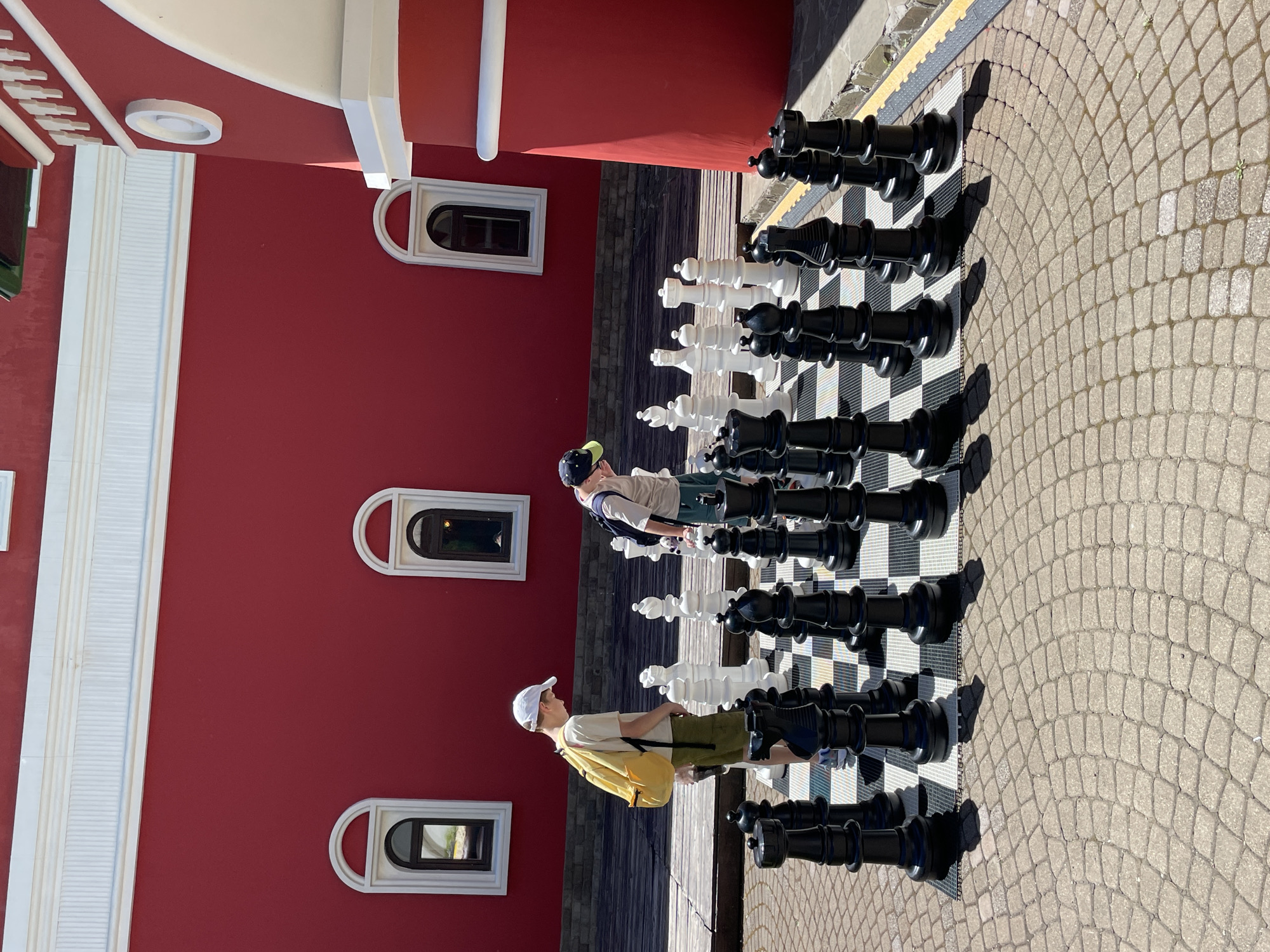 Two young males are playing a game of giant chess outdoors on a paved surface in front of a red building with white trim and arched windows. The male on the left, wearing a white baseball cap, a light-colored t-shirt, and green shorts, has a yellow backpack slung over his right shoulder. He is looking down at the chessboard. The male on the right, wearing a dark baseball cap and a light-colored t-shirt with green shorts, is also looking down at the chessboard. The chess pieces are large, with black and white sets. The environment appears to be a public space, possibly a park or plaza, with wooden decking visible behind the chess board. The building's architecture suggests it might be a historical or culturally significant structure. The location is Estosadok, Russia.