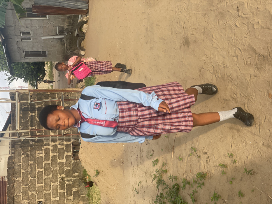 The image depicts two female students walking on a dirt path in Mosafejaw, Nigeria. The foreground features an adolescent female, with dark skin and short dark hair, wearing a light blue long-sleeved school shirt with a crest on the left pocket, a red tie, a red and white plaid skirt, light grey socks, and black lace-up shoes. She carries a black backpack and walks with her head slightly down, looking forward. To her right and further back, a younger female child, also with dark skin and dark hair, wears a light pink short-sleeved top, a matching plaid skirt, and black shoes. She carries a bright pink bag in her left hand and looks towards the viewer.
The environment includes a rough concrete block wall on the left, with a pile of sand or dirt and sparse green vegetation. A dark-feathered fowl stands on the dirt pile. Behind this wall, a metal structure is visible. On the right, a grey building with small windows stands, with discarded tires and corrugated metal sheets near its base. In the far background, a faint figure walks on a distant path, and a telecommunications mast is visible against the sky. Green foliage and trees are also present in the background. The ground is a light-colored sandy dirt path with visible footprints. The scene is illuminated by natural daylight.