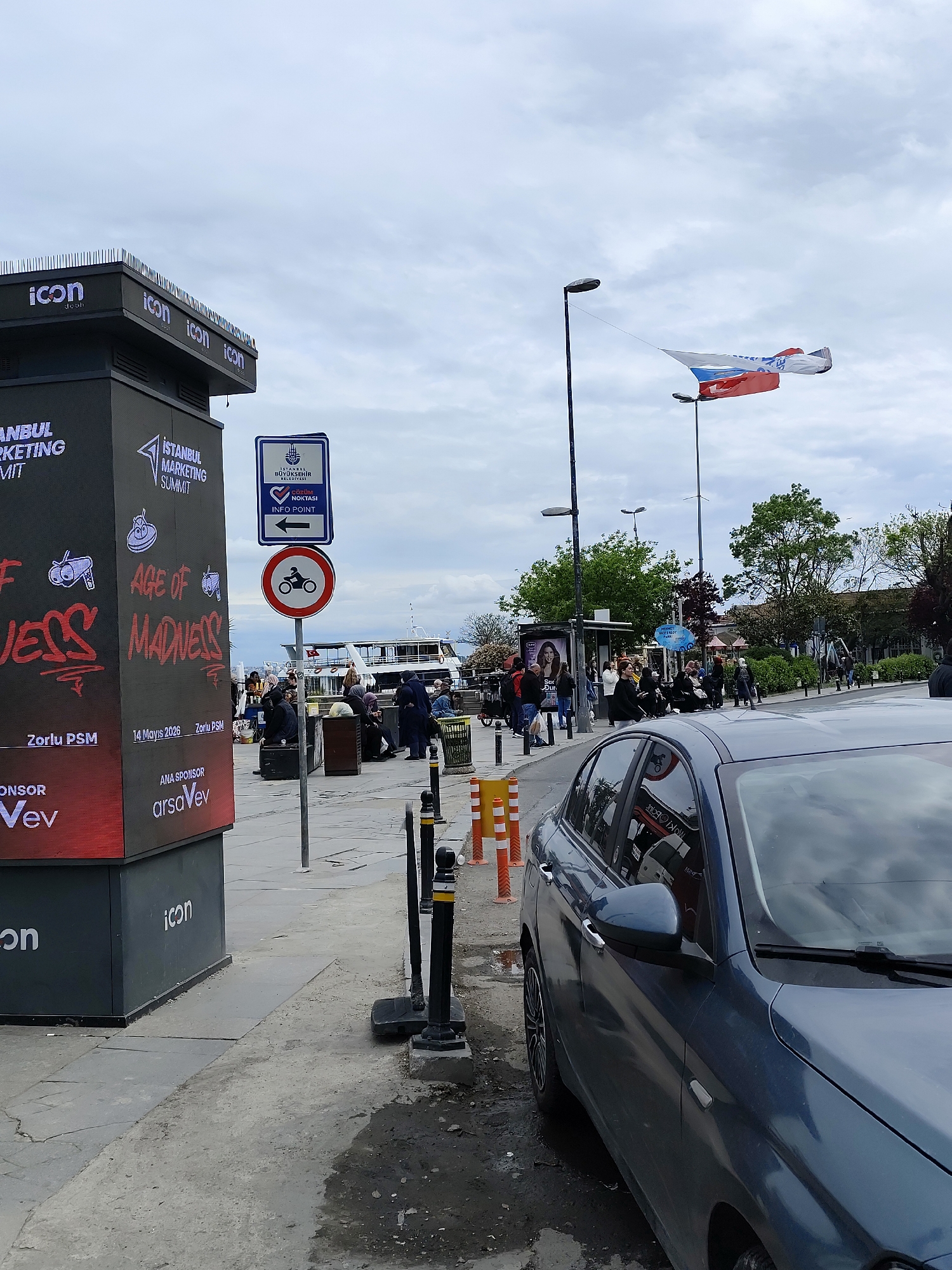 The image shows a street scene, likely in Istanbul, Turkey, as indicated by the "ISTANBUL MARKETING SUMMIT" sign. The weather appears overcast, with a cloudy sky. The foreground is dominated by a dark grey car parked on the side of the road. To the left of the car is a large, black digital display board promoting the "ISTANBUL MARKETING SUMMIT" event, which includes the text "AGE OF MADNESS". Adjacent to the display board is a sign indicating "INFO POINT" with a no-motorcycle traffic sign.
In the background, people are gathered along the waterfront, possibly waiting for a ferry, as a boat can be seen in the distance. There are trees and buildings lining the waterfront. A large, dual-colored flag is visible overhead, possibly a promotional flag, strung between two light posts. Other street furniture such as orange and white bollards and dark posts with square bases are also visible.
