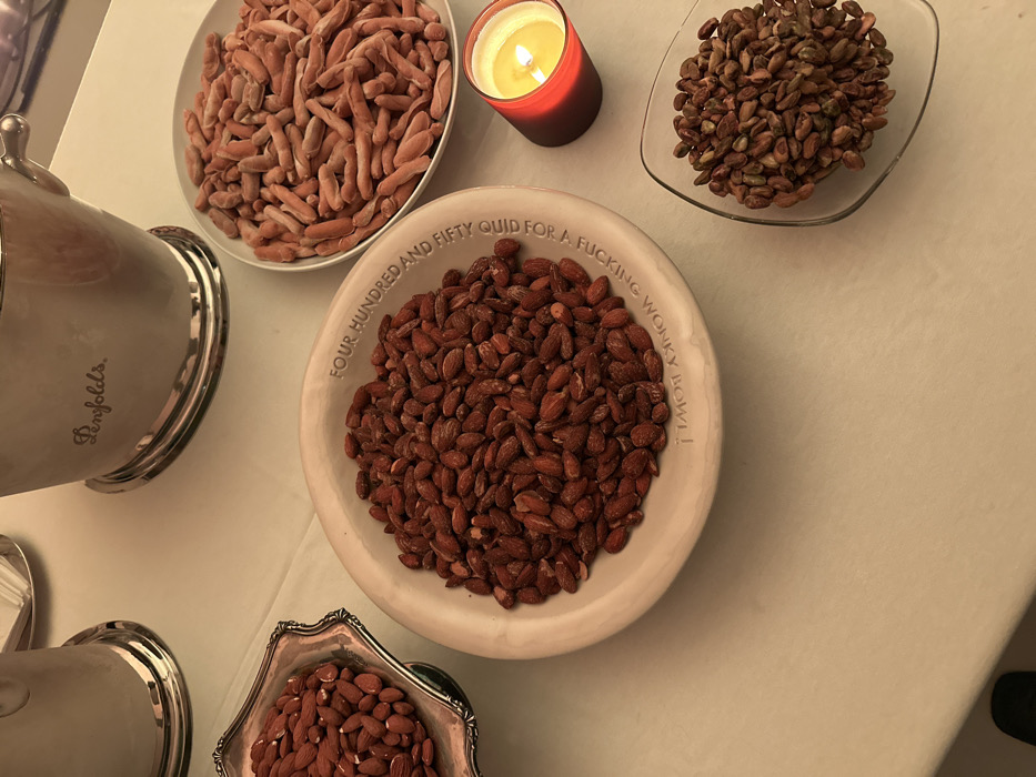 The image showcases several bowls filled with food. There are bowls of reddish-brown nuts, and a bowl of small, sausage-shaped items. A lit candle sits near one of the nut bowls. A large, silver container, labeled "Penfolds," is partially visible on the left. The central bowl is inscribed with the phrase "FOUR HUNDRED AND FIFTY QUID FOR A FUCKING WONKY BOWL!"