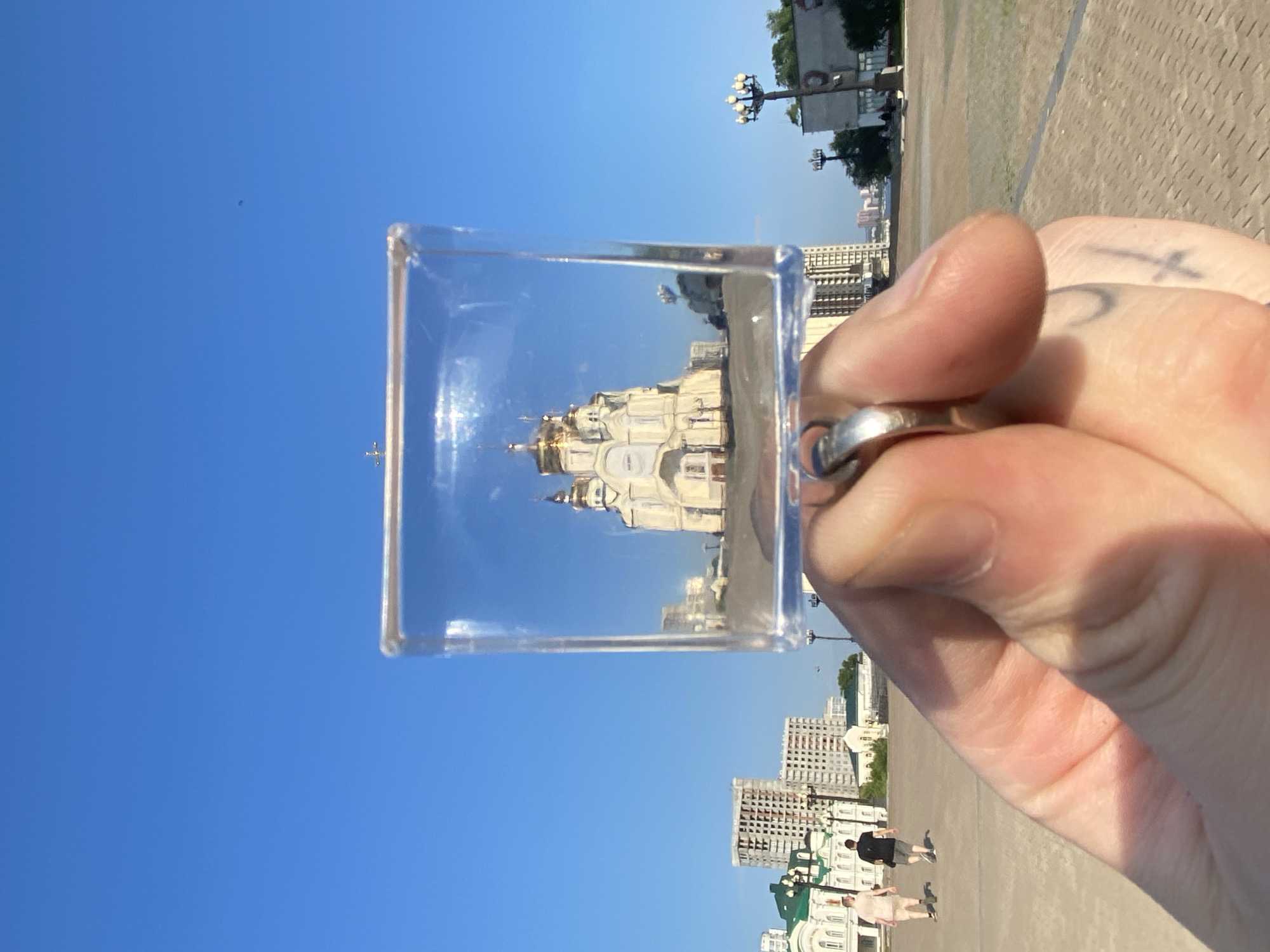 A person's hand, wearing a silver ring, holds a clear cubic glass object in front of a city scene. The glass cube contains a reflected image of the Transfiguration Cathedral in Khabarovsk, Russia, with its golden domes and white facade. The background shows an open paved area, a street with lampposts and trees, and buildings under construction. Two individuals are walking in the distance across the paved area. The sky is clear and blue.