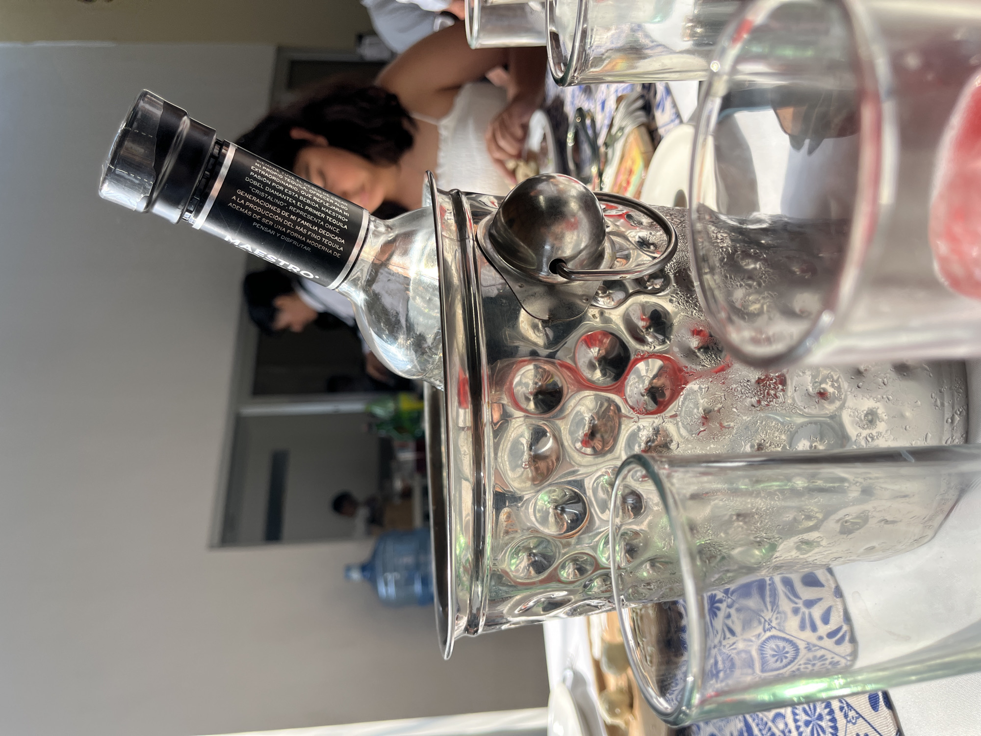 The image shows a bottle of Maestro Dobel Diamante tequila in an ice bucket, with several empty glasses around it. In the background, a person is visible at a table. Further back, a water cooler is visible. The location is San Bernardino Tlaxcalancingo, Mexico.