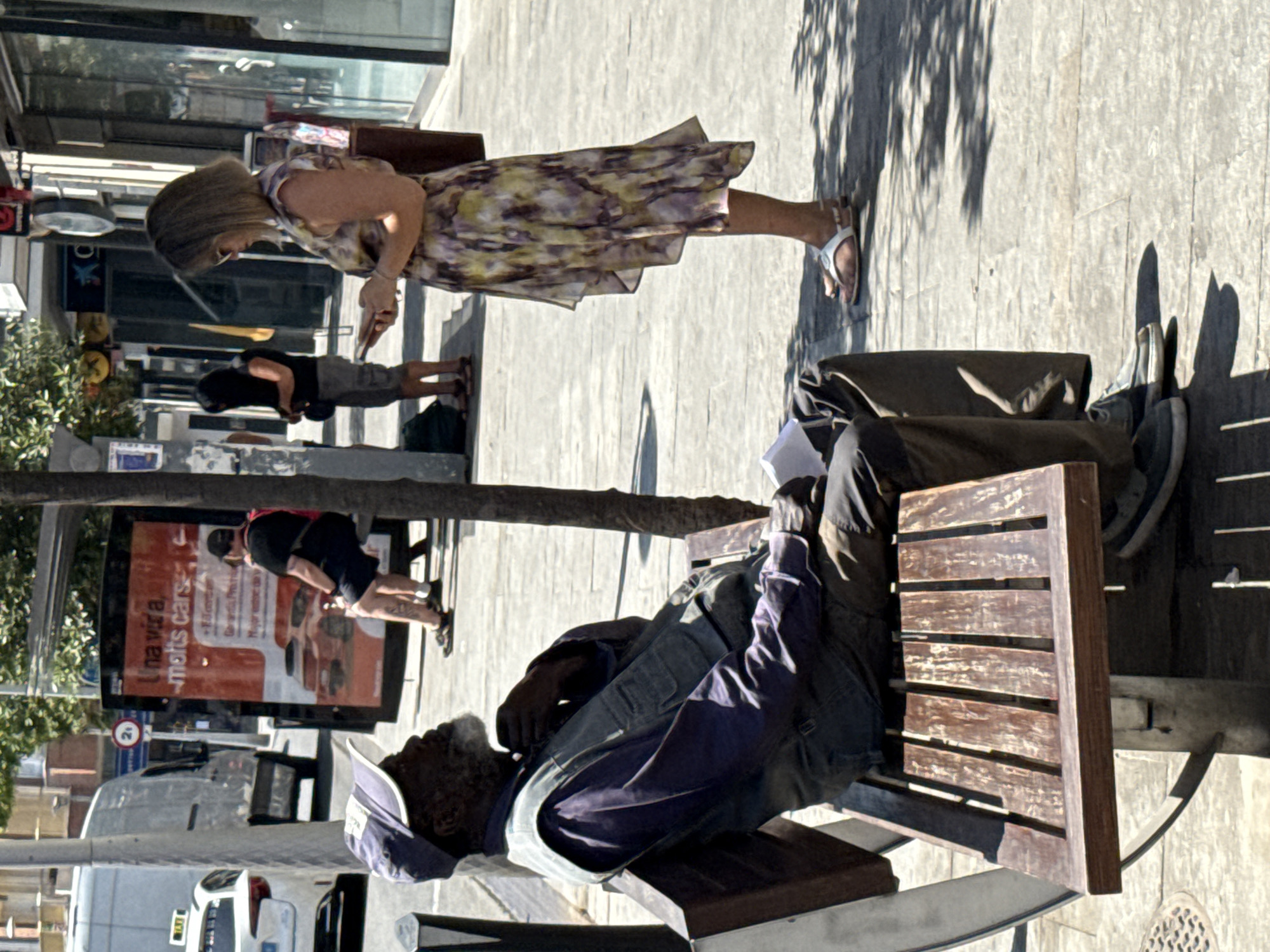 A man wearing a blue baseball cap, a purple long-sleeved shirt, and a grey vest sits on a wooden slatted bench on a sunny day. He has a grey beard and is looking upwards with his right hand near his face. A piece of white paper is visible on his lap. To the right of the bench, a woman in a light-colored, floral-patterned dress and white sandals is standing and looking at her phone. Behind the man and bench is a tree. In the background, a large advertisement board is visible, along with buildings and people walking on a sidewalk. A white van and a taxi are parked on a street to the left of the frame. The scene appears to be a public area in the city of Blanes, Spain, likely a street or a promenade.