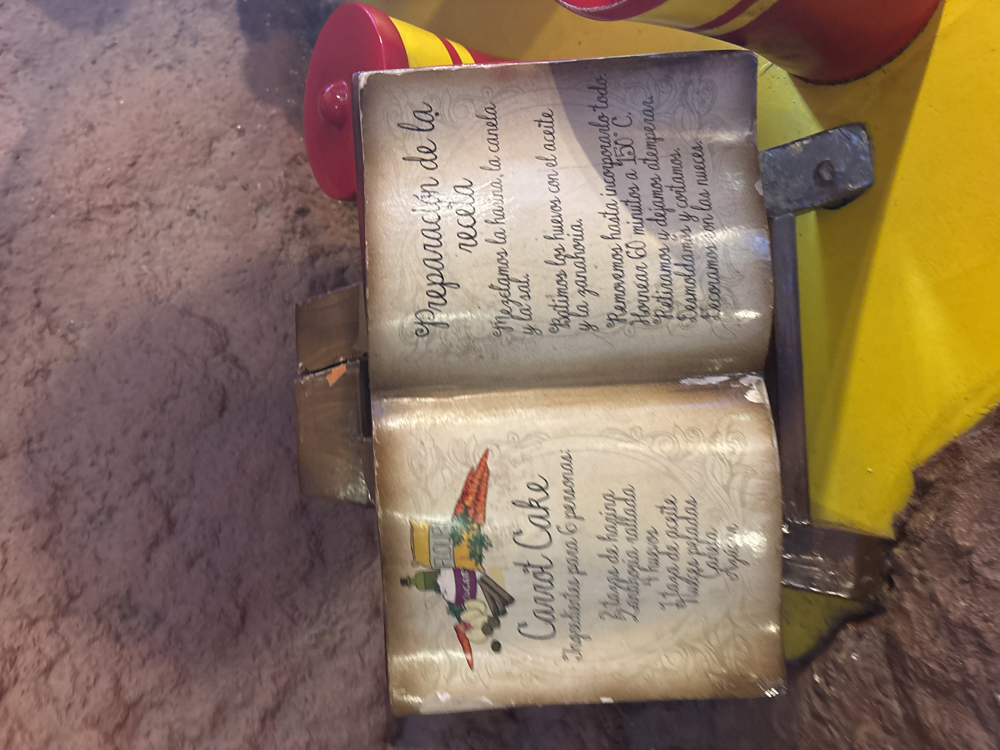 This image depicts a wooden carving resembling an open recipe book, situated indoors. The book's pages feature recipes in Spanish. The left page has an illustration of a Carrot Cake, along with a list of ingredients intended for six people, including flour, grated carrots, eggs, oil, peeled nuts, and cinnamon. The right page contains instructions titled "Preparación de la receta" (Preparation of the recipe), detailing how to mix the ingredients, bake at 150°C for 60 minutes, and decorate with nuts. The carving appears to be part of a larger decorative structure, as suggested by the visible portion of a yellow and red object in the background. The exact location in San Martín de la Vega, Spain, is impossible to determine from the image alone.