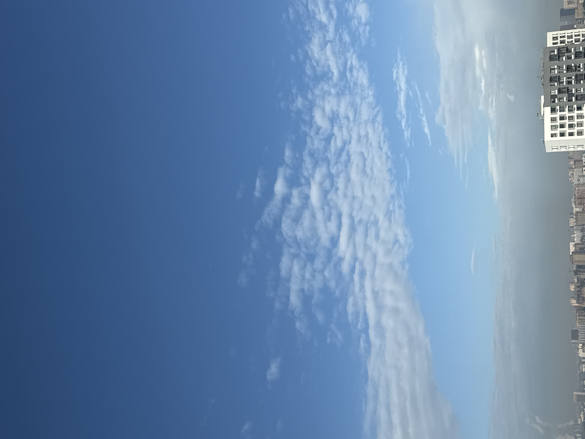 The image shows a clear blue sky with scattered cirrocumulus clouds over Lyubertsy, Russia. The right side of the image shows a dense cityscape, including a tall, modern, white building with a grid-like facade. The overall environment suggests a bright, sunny day in an urban area.