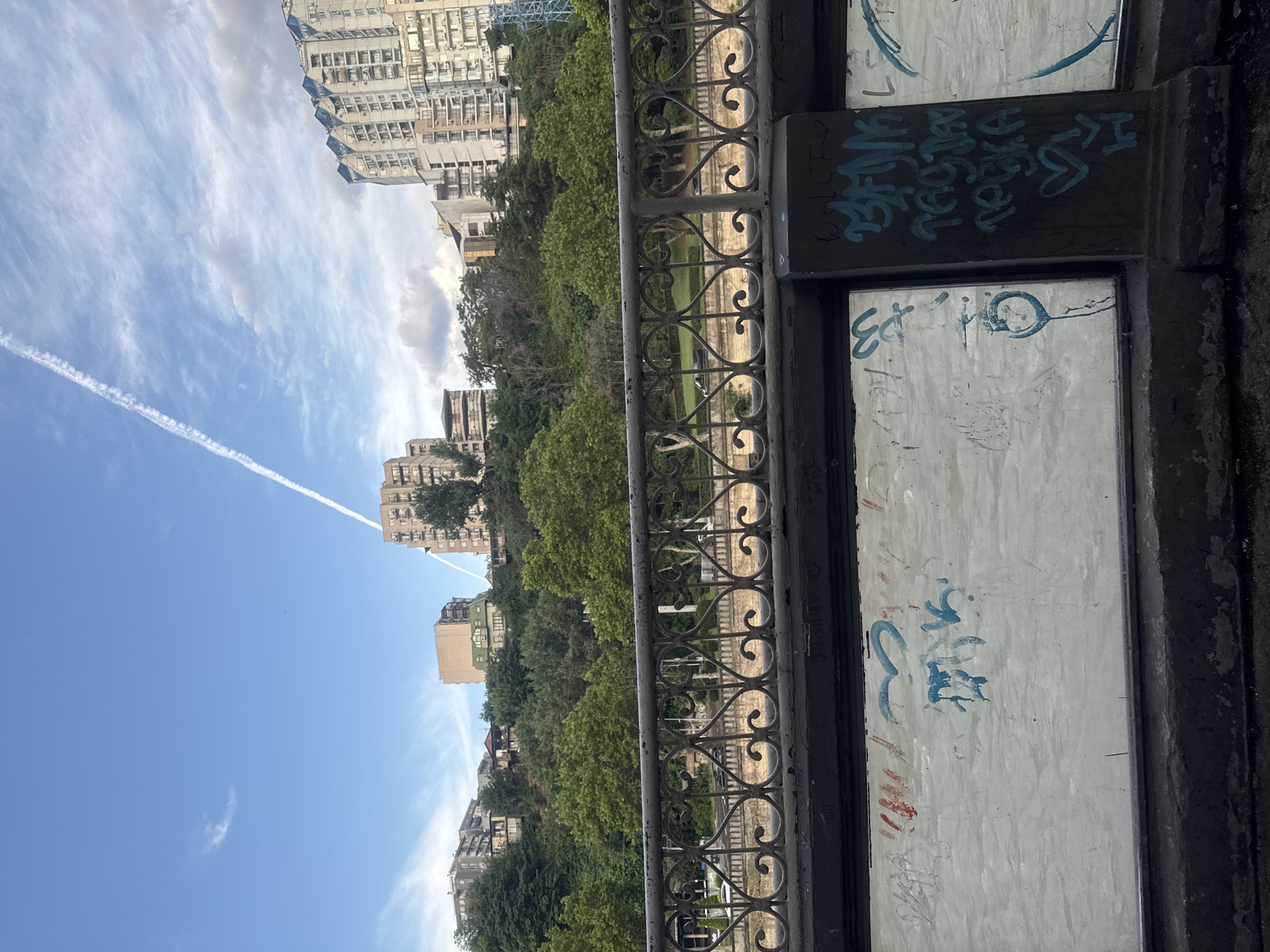 A view from a bridge overlooking a river in Tbilisi, Georgia, showcases a dense urban landscape. In the foreground, a dark, graffiti-covered railing and wall obscure the view. Beyond, a lush green hillside is densely populated with trees. Several multi-story buildings are nestled among the trees, with taller, modern apartment buildings dominating the skyline further in the background. The sky above is a clear blue with scattered clouds and a prominent contrail from an airplane. The lighting suggests it is daytime, likely late afternoon or early evening given the quality of light.