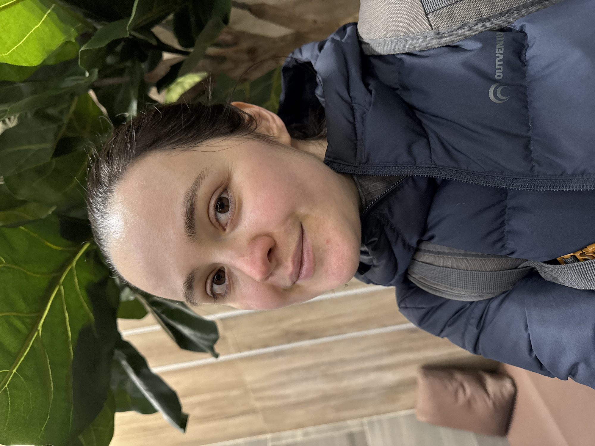 A woman with dark hair and a navy blue "OUTVENTURE" jacket is looking at the camera. She is standing near a large green plant, possibly a fiddle-leaf fig, in a room with light brown, horizontally-patterned flooring. A brown chair or cushion is partially visible in the lower right corner. The location is unknown and cannot be determined as Izhevsk, Russia, from the image alone.