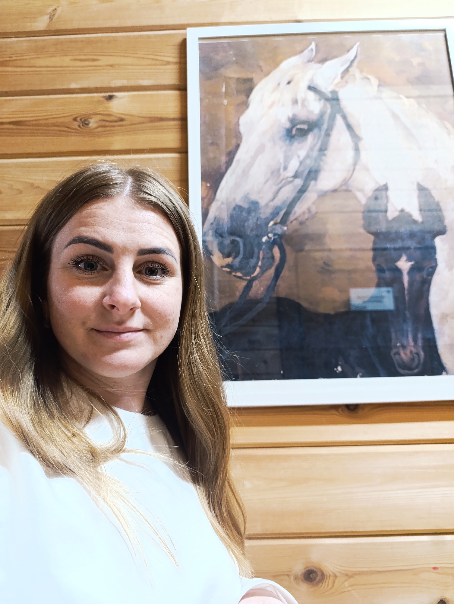 A woman is standing in front of a wooden wall on which hangs a framed painting of two horses. The woman has light brown hair and is wearing a white top. The painting shows the head and upper body of a white horse with a black bridle and the head of a dark horse in the background. The environment suggests an interior setting, possibly a home or gallery. The location is unknown.