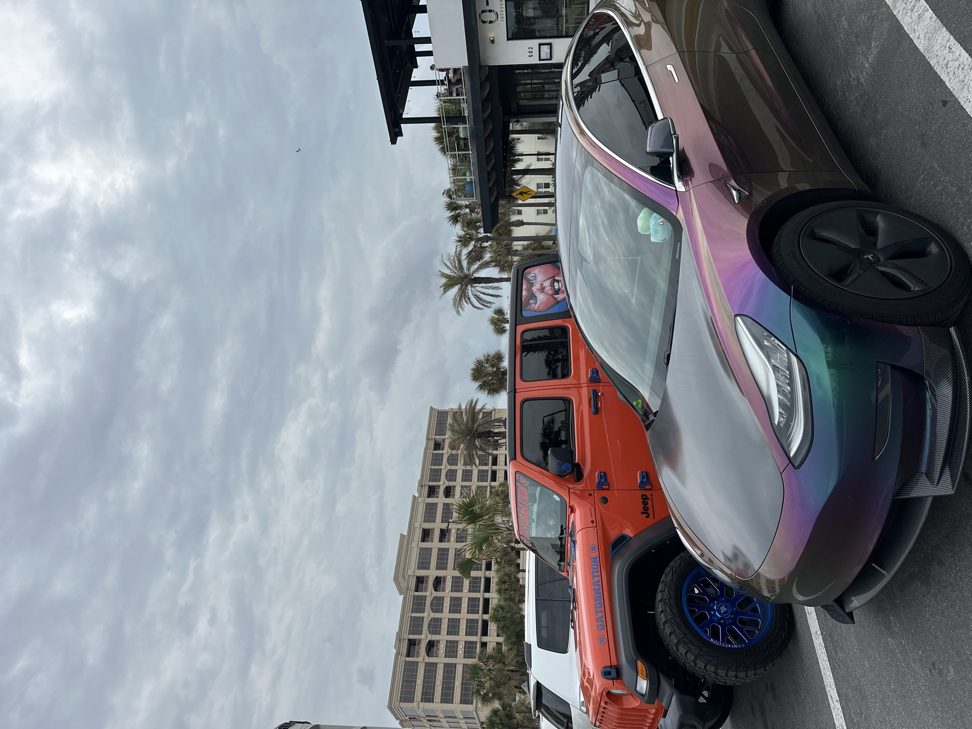 The photograph depicts a street scene in Jacksonville Beach, Florida, United States. The camera is positioned at street level, capturing a row of parked vehicles. Prominently featured are a custom-wrapped Tesla with iridescent paint, a vibrant orange Jeep Wrangler with blue wheels, and a white pickup truck. A multi-story building with numerous windows is visible in the background, adjacent to palm trees. A restaurant or commercial building with architectural features, including an awning, is partially visible at the top. The sky is overcast with a cloudy atmosphere, and the time of day appears to be during daylight hours.