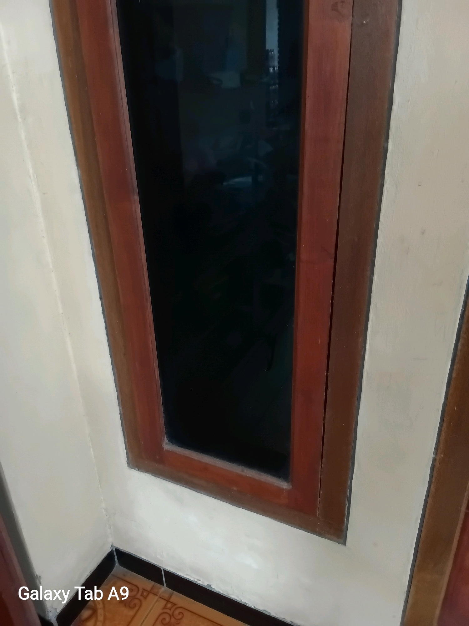 A close-up, vertical shot shows the corner of a room with a tall, narrow window framed by dark brown wood. The window pane is dark, reflecting what appears to be dimly lit interior space. The wall to the left of the window is painted a light, creamy color, showing some texture and slight discoloration at the bottom corner. A dark tile baseboard runs along the floor. The bottom left corner of the image displays a watermark that reads "Galaxy Tab A9". The scene suggests an indoor environment, possibly a residential or office space, with a focus on architectural details.