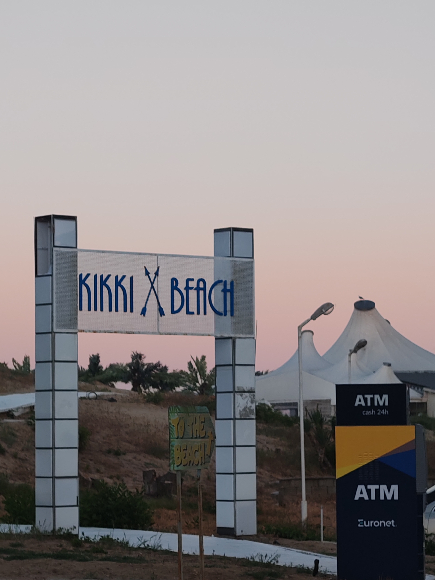 The image shows the entrance to Kikki Beach in Nesebar, Bulgaria. The sign reads "KIKKI BEACH" with crossed arrows instead of an "X." A smaller, weathered wooden sign says "TO THE BEACH!" An ATM sign for Euronet is visible on the right. In the background, there is a large white tent structure. The sky is light pink/orange, indicating either sunrise or sunset. The ground is dry and sparsely covered with vegetation.