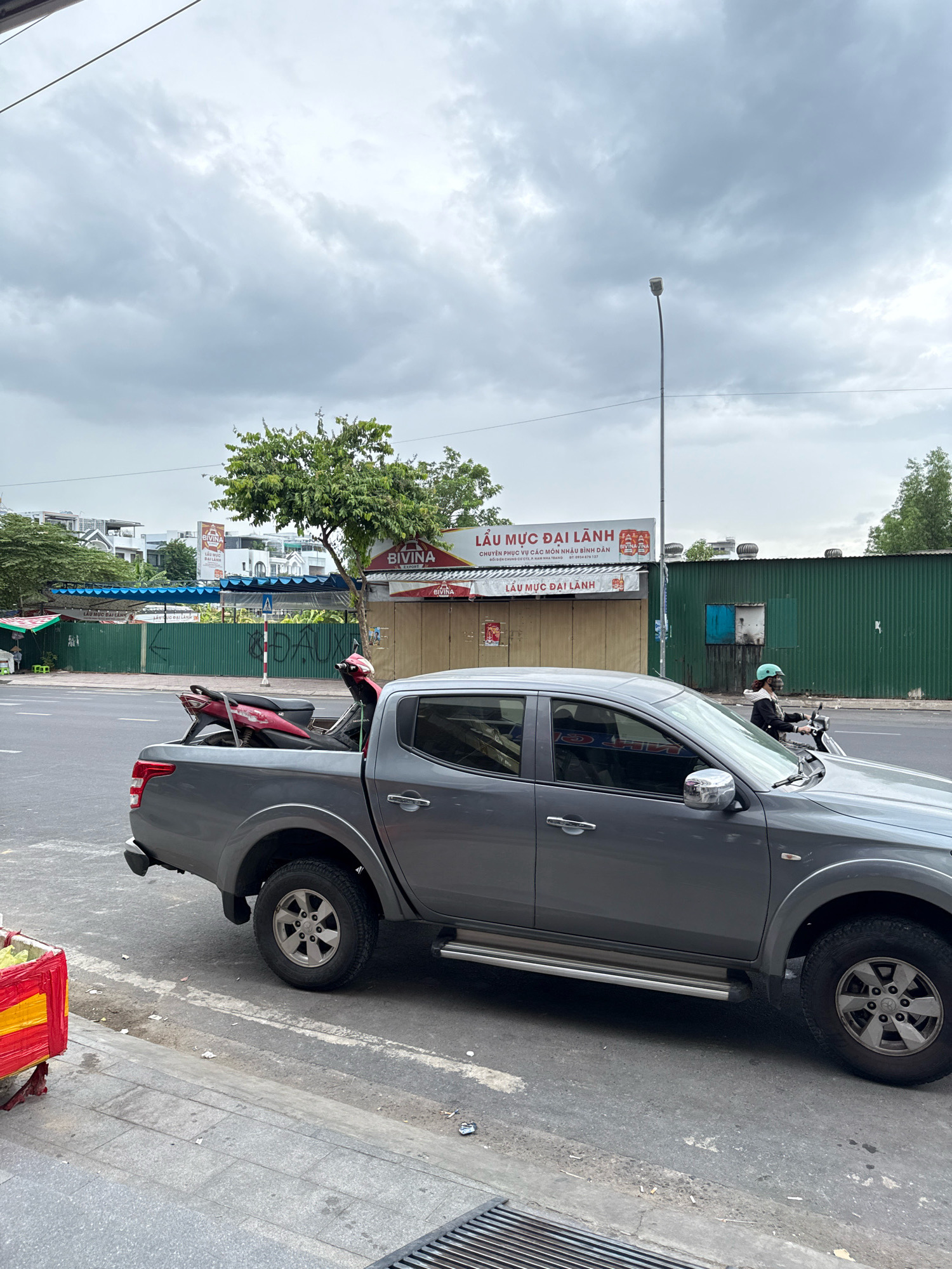 A gray pickup truck is parked on the side of a street in Nha Trang, Vietnam, with a red scooter loaded in its bed. The truck is a modern, four-door model with chrome accents on the door handles and side mirrors. The scooter appears to be a relatively small, lightweight model, possibly a Honda. The street is paved and appears to be in a commercial or residential area, with buildings and trees visible in the background. The sky is overcast, suggesting a cloudy day. A person on a motorcycle is seen passing the truck on the right side of the frame. In the background, there are signs in Vietnamese, one of which advertises "LẨU MỰC ĐẠI LÀNH," which translates to "Big Squid Hot Pot." The overall scene depicts a typical street view in a Vietnamese city, with a mix of vehicles, businesses, and everyday life.