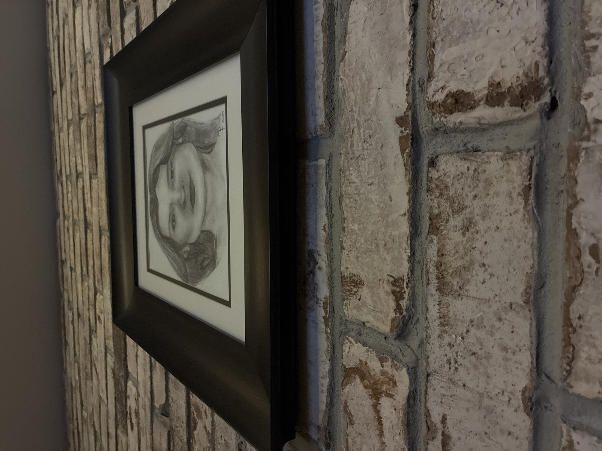 A close-up, angled photograph captures a framed pencil drawing displayed on a textured wall. The central subject is a grayscale portrait, likely of a young woman or girl, depicted with a gentle smile and her head tilted slightly. Her hair appears to be swept back or to one side. The drawing is encased in a dark-colored, possibly black or very dark brown, frame with a white mat board.
The wall exhibits distinct textures. To the left of the frame, there's a section covered with narrow, vertically oriented, light-colored bricks or tiles. To the right of the frame, the wall is composed of larger, irregularly shaped bricks or stones that are primarily light-colored, possibly painted white or light grey. These larger bricks show significant texture and wear, with hints of a darker, reddish-brown material beneath the surface, separated by visible, pronounced dark grey mortar lines.
The lighting is somewhat subdued, creating soft shadows and highlighting the various surface textures. In the upper right corner of the pencil drawing, faint, handwritten text is discernible, including what appears to be a signature or initials and the year "2019". The image portrays a static, indoor scene focused on the artwork and its immediate surroundings.
