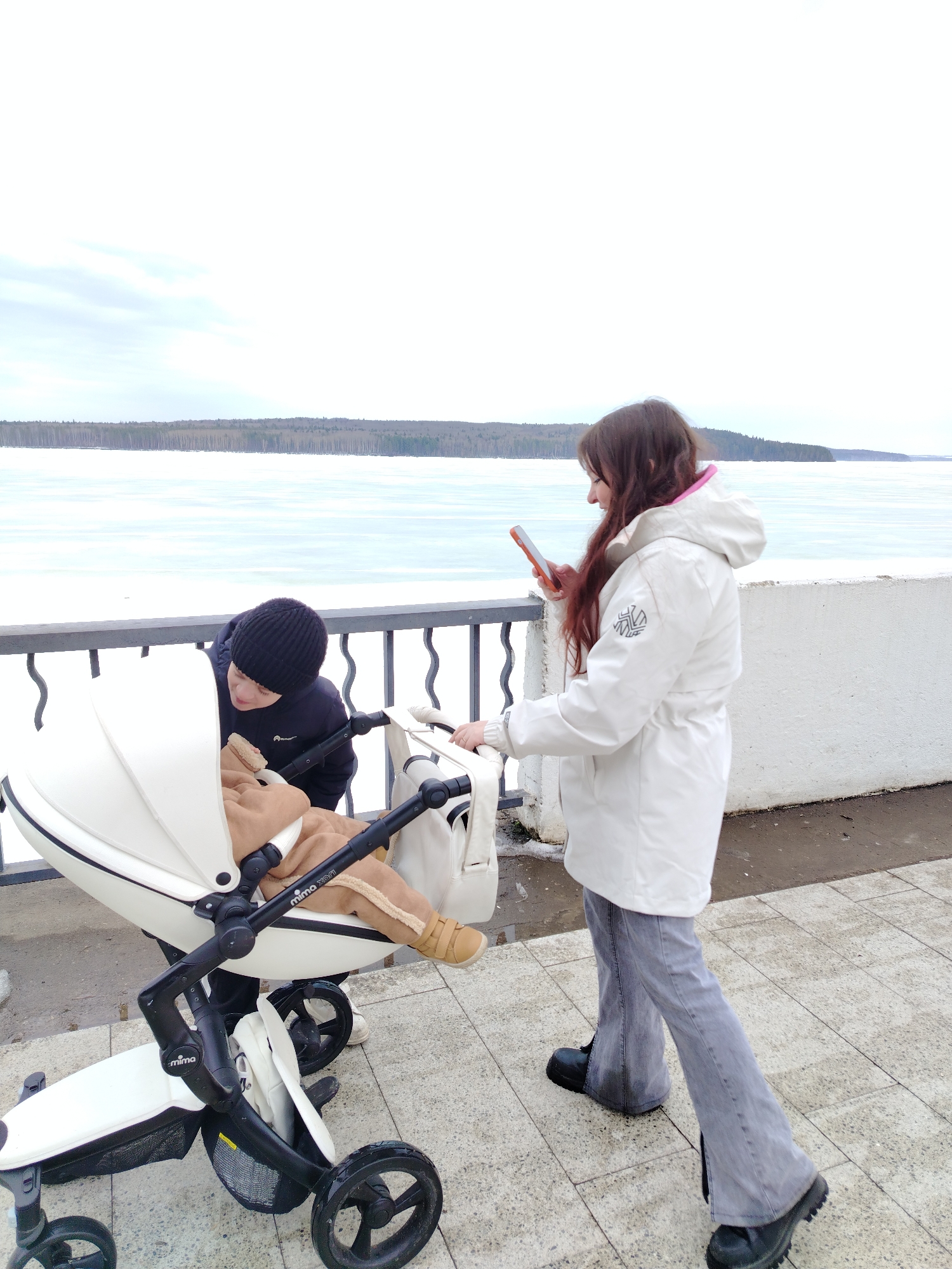 The image depicts a scene in Chaikovsky, Russia, along a paved walkway beside a body of water, likely the Kama River given the location. The water appears to be partially frozen or covered in ice and snow.
Two individuals are present: a young woman and a younger person, potentially a child, wearing a black hat and dark clothing, bending over a white stroller. Inside the stroller is an infant, wearing tan clothing and boots, and covered by a tan blanket. The stroller is a 'mima' model.
The young woman is holding a mobile phone, possibly taking a picture, and is dressed in a white hooded jacket, gray jeans, and black boots. The sky is overcast. A white concrete barrier is visible, with a metal railing behind it.