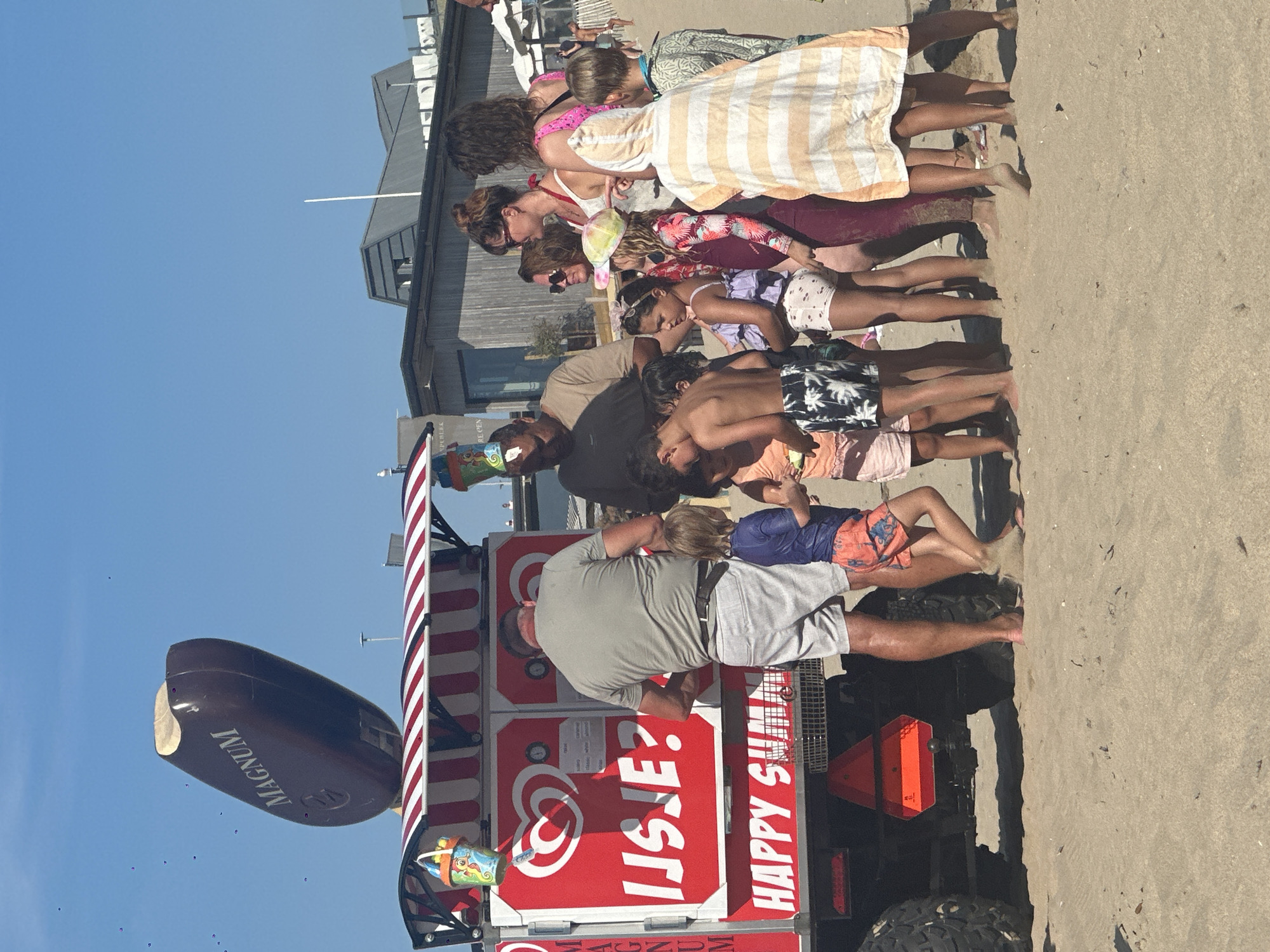 The image depicts a sunny day on a sandy beach in Zandvoort, Netherlands. A red ice cream truck, adorned with a large inflatable Magnum ice cream cone on top and the text "IJsje?" (Dutch for "Ice cream?") and "HAPPY SUMMER" on its side, is the central focus. A group of people, including several children and adults, are gathered around the truck, presumably purchasing or waiting for ice cream. The children are dressed in swimwear, while the adults wear casual beach attire. In the background, modern beachside buildings and umbrellas can be seen. The sky is clear and blue.