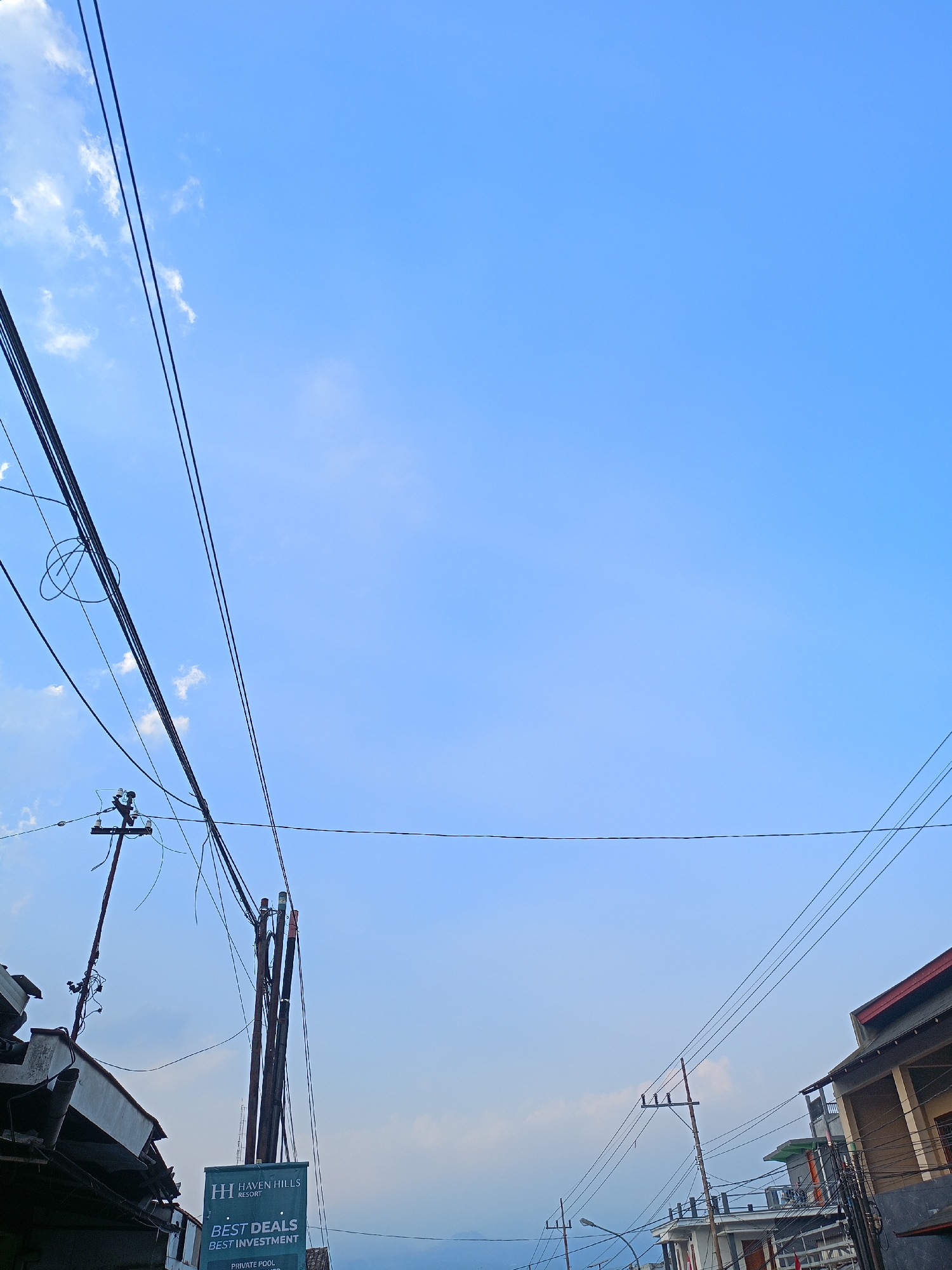 The image depicts a clear blue sky with some wispy clouds, with numerous power lines crisscrossing the frame. In the lower left, part of a building with exposed rebar and incomplete construction is visible. A utility pole with electrical equipment and wires is also present. To the right of the utility pole, a tall, multi-stemmed structure with what appear to be colored tubes or conduits attached is standing. A green banner hangs below this structure, bearing the logo "H HAVEN HILLS RESORT" and text that reads "BEST DEALS BEST INVESTMENT". Further to the right, a multi-story building with balconies and a red-tiled roof is partially visible. Another utility pole with wires is seen in the middle ground. The overall scene suggests an urban or semi-urban environment with ongoing infrastructure and construction, likely in a location with a view of natural landscapes, given the resort advertisement. The setting is the city of Batu, Indonesia, known for its cool climate and tourist attractions.